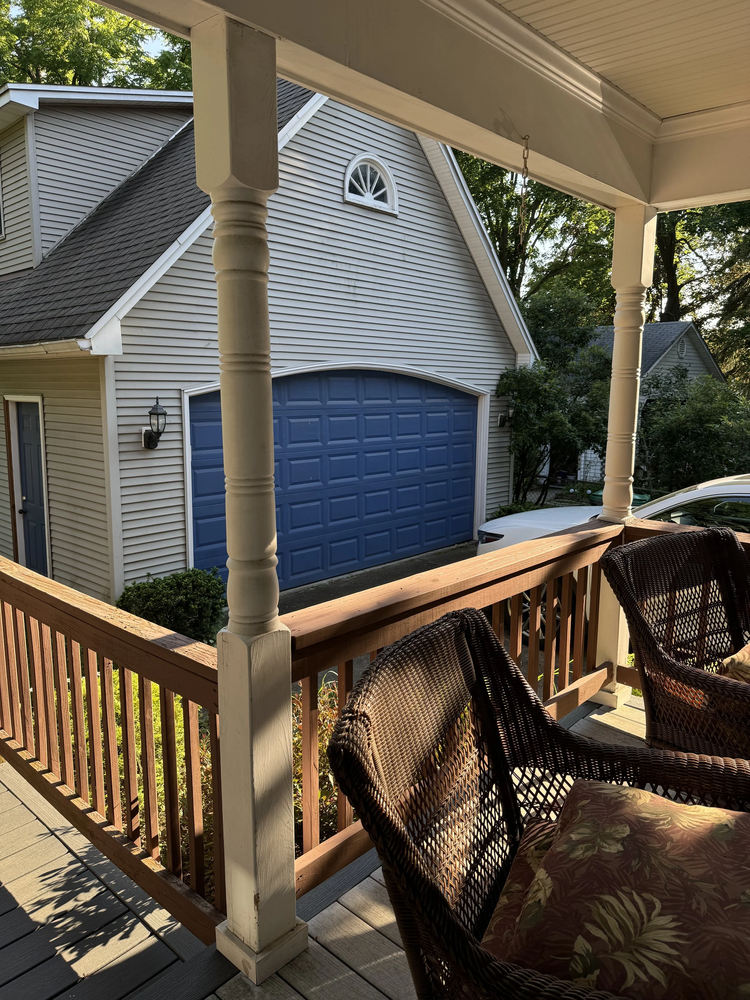 View from a porch showing wicker chairs with a floral cushion, wooden railing, and beige columns. In the background, residential houses with vinyl siding, a garage with a blue door, and trees are visible.