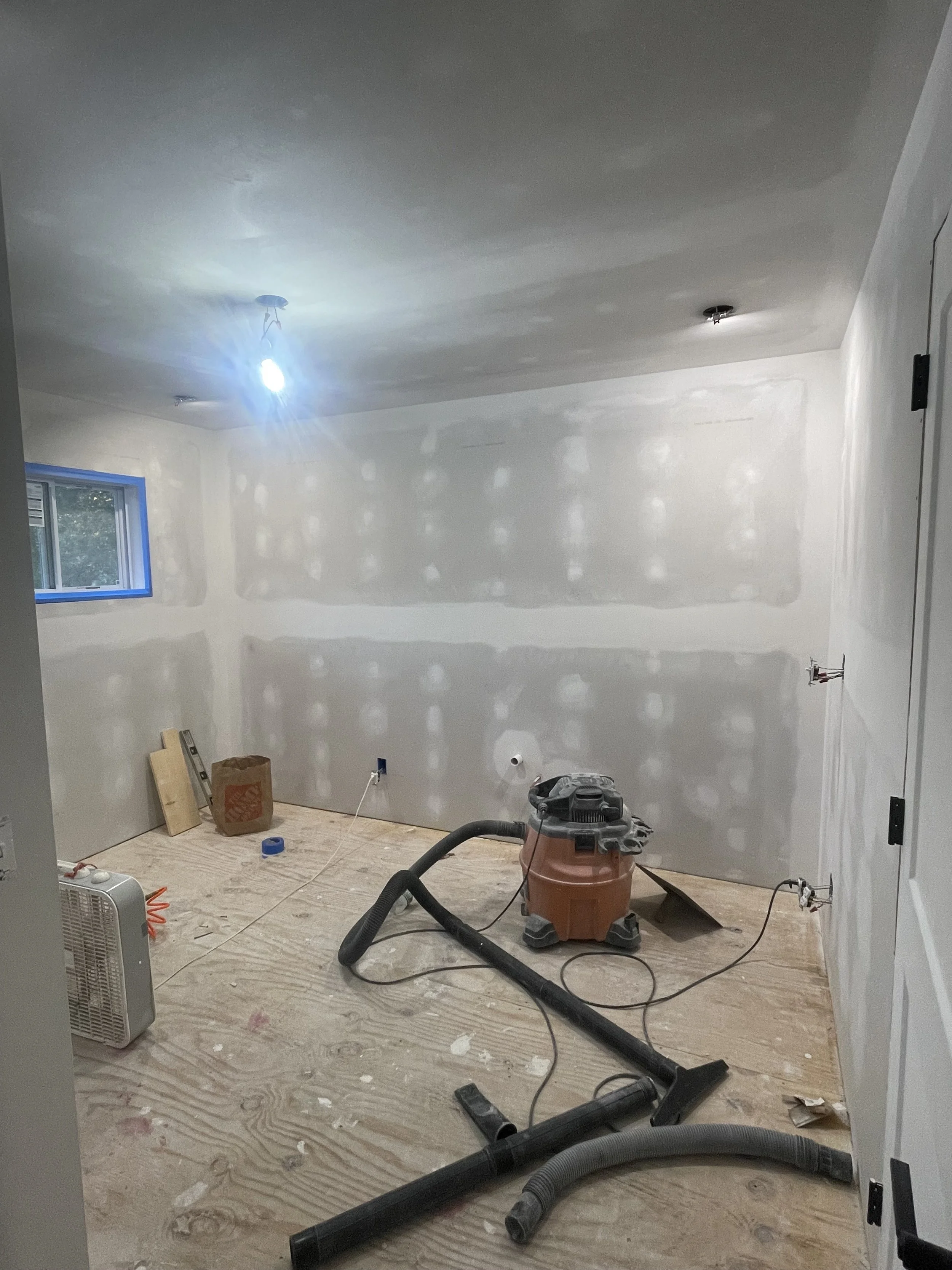Room under construction with drywall on walls, construction tools, vacuum, and plywood on floor.