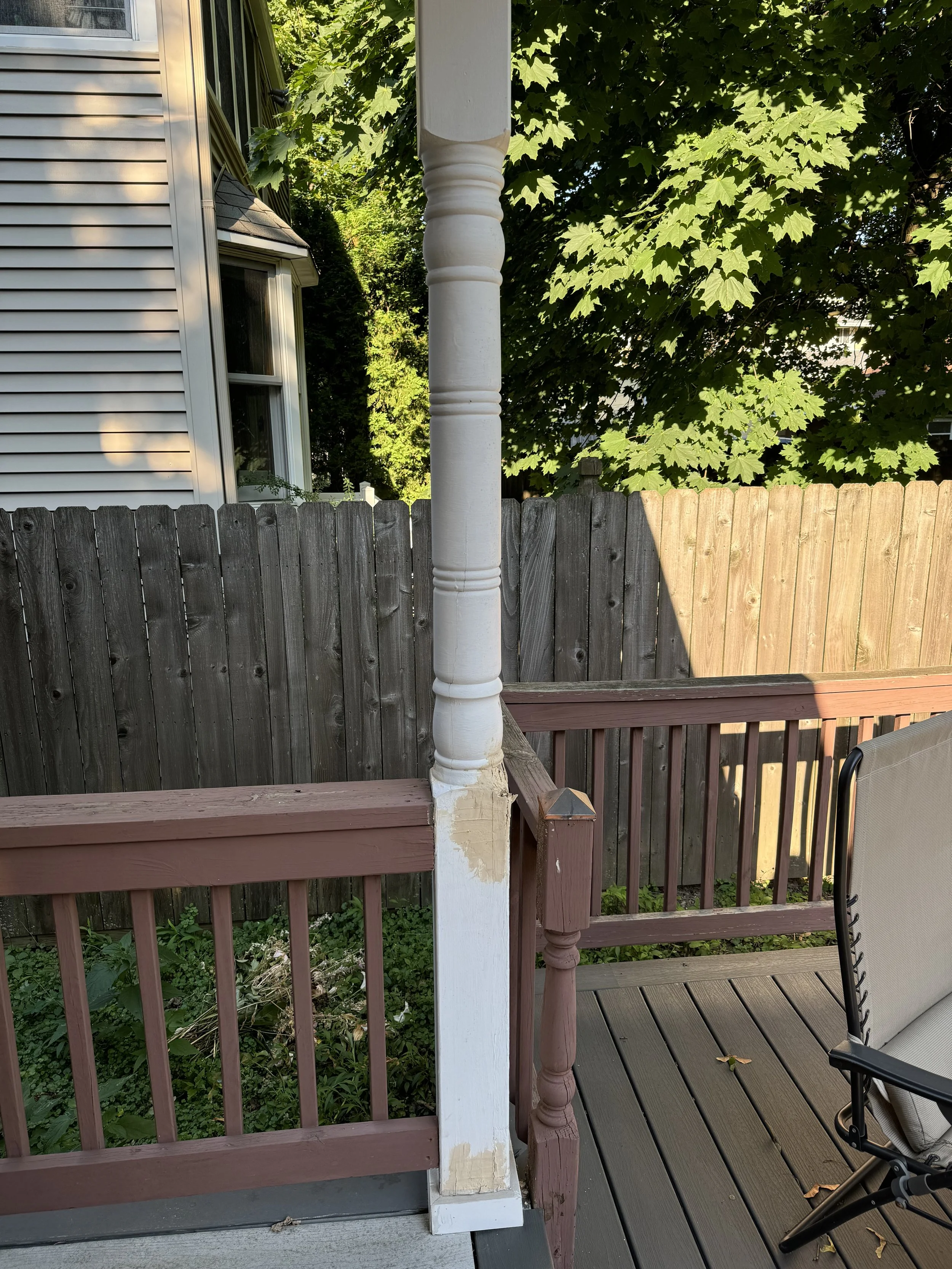 A backyard deck with wooden railing and a white and pink support post. There is a green tree with large leaves in the background and a wooden fence. A beige outdoor chair is partially visible on the right side of the image.