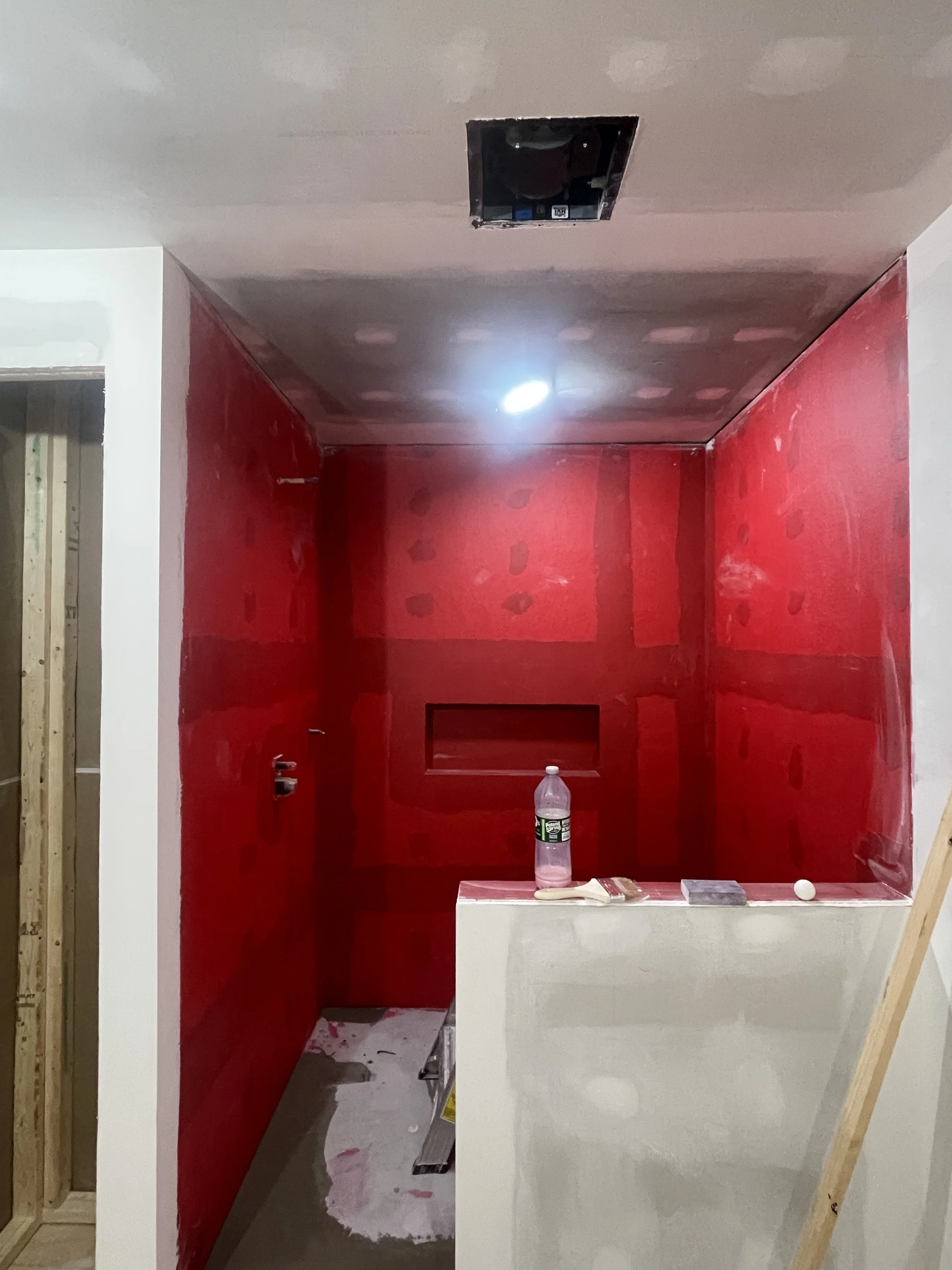 A bathroom under construction with red drywall on the walls, a small recessed shelf, and an open ceiling vent. There are construction materials and tools on a white half-wall in the foreground.