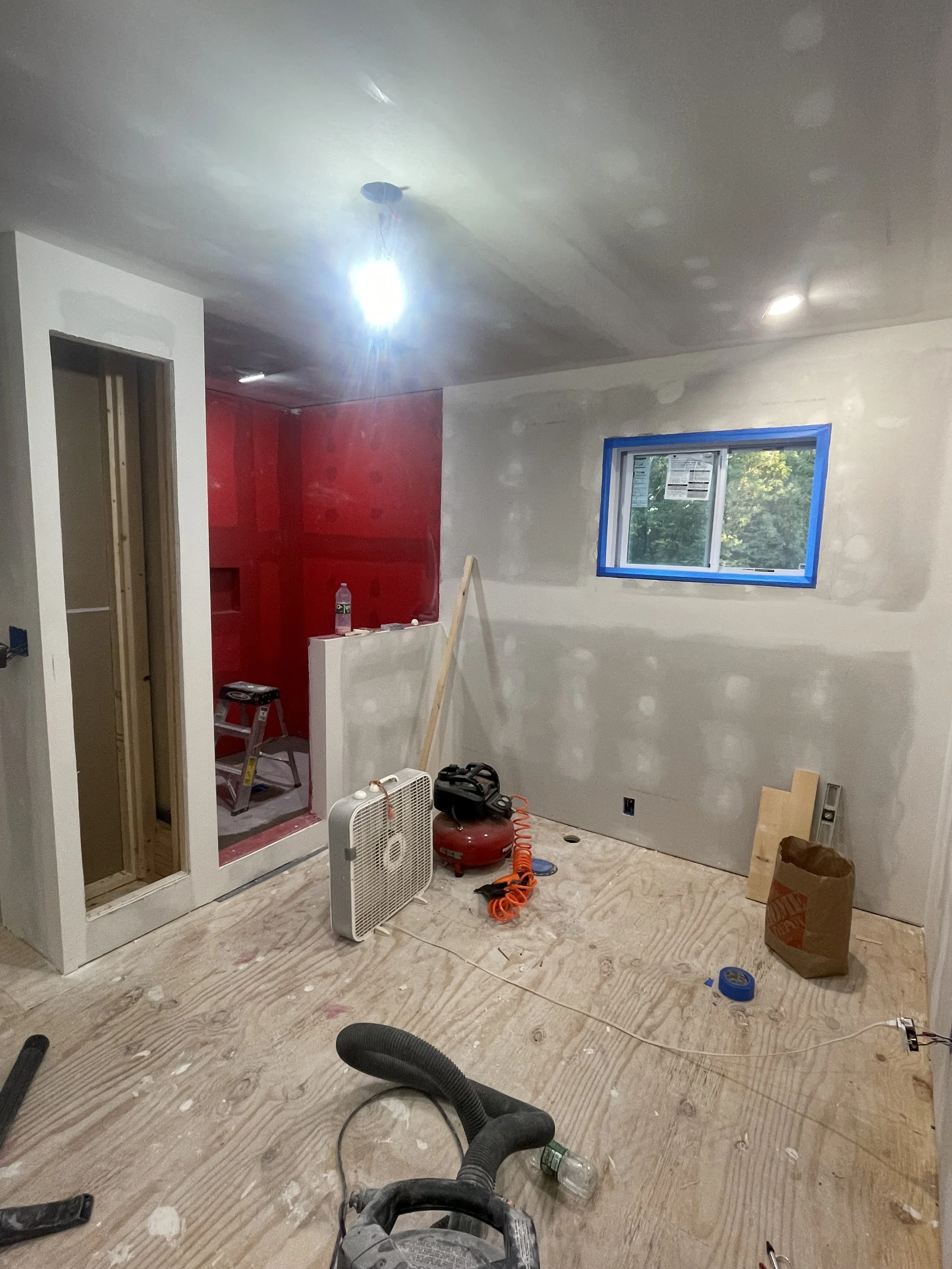 Interior of a room under construction with unfinished drywall on the walls, a window with blue tape around its edges, construction tools and materials on the floor, and an unfinished closet or doorway.
