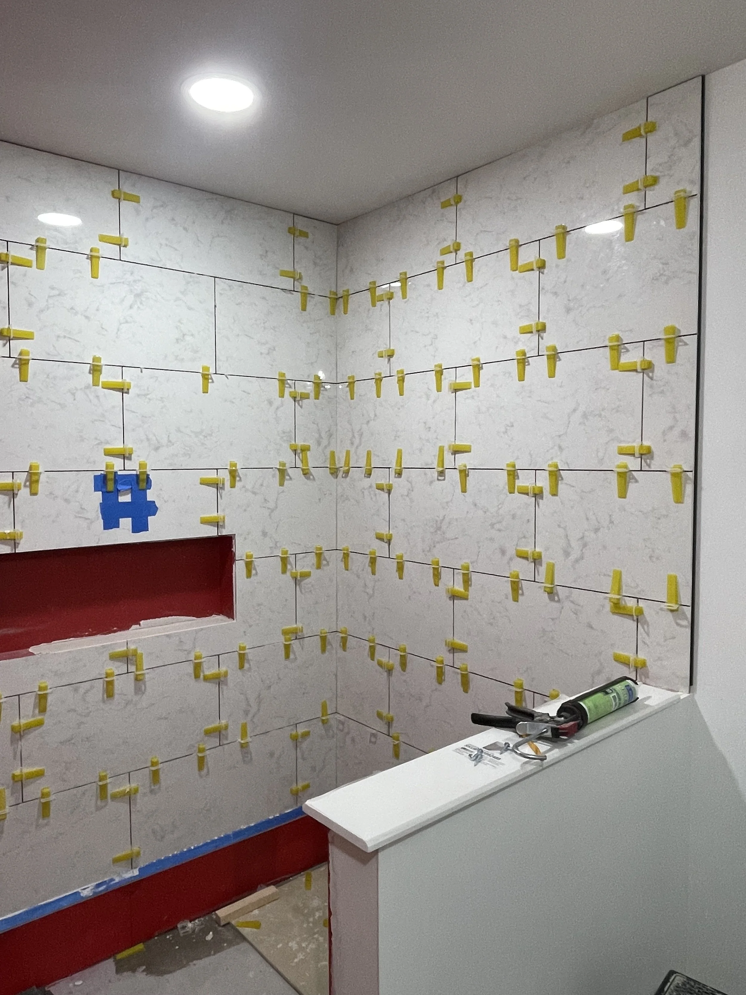 A corner of a room under construction with white marble wall tiles. Multiple yellow tile spacers are arranged between the tiles, with a few blue spacers shaped like a character hanging on the wall. Tools and materials are on a white ledge in the fore