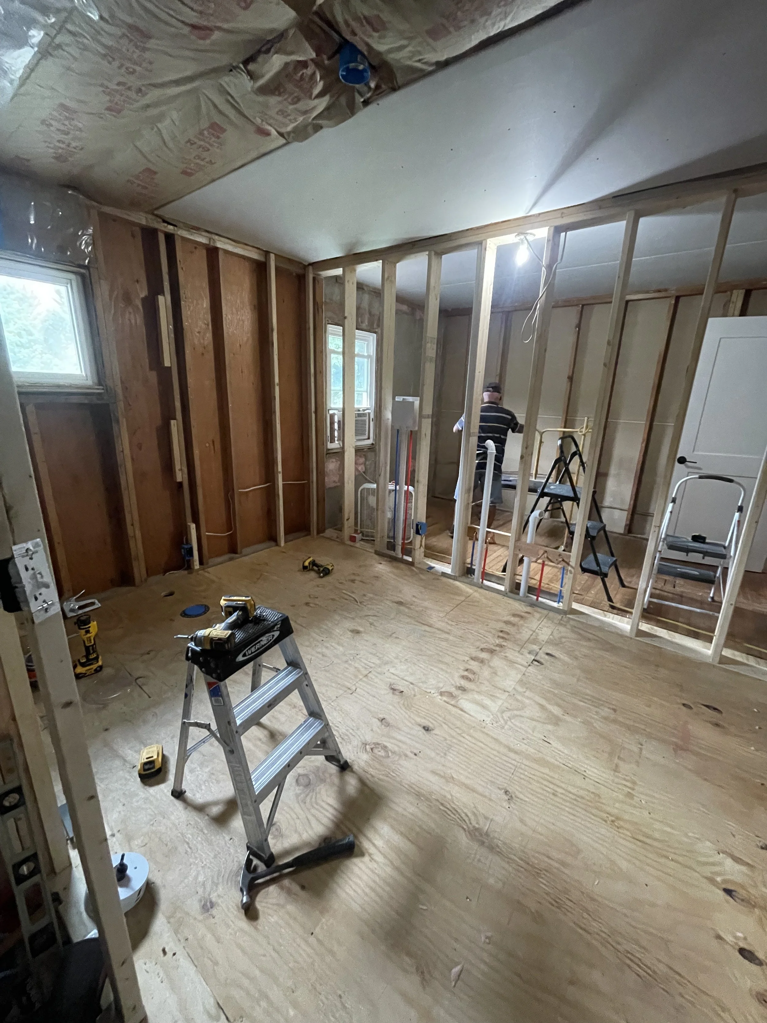 Interior of a room under renovation with exposed wooden framing, tools on the floor, and a person working in the background.