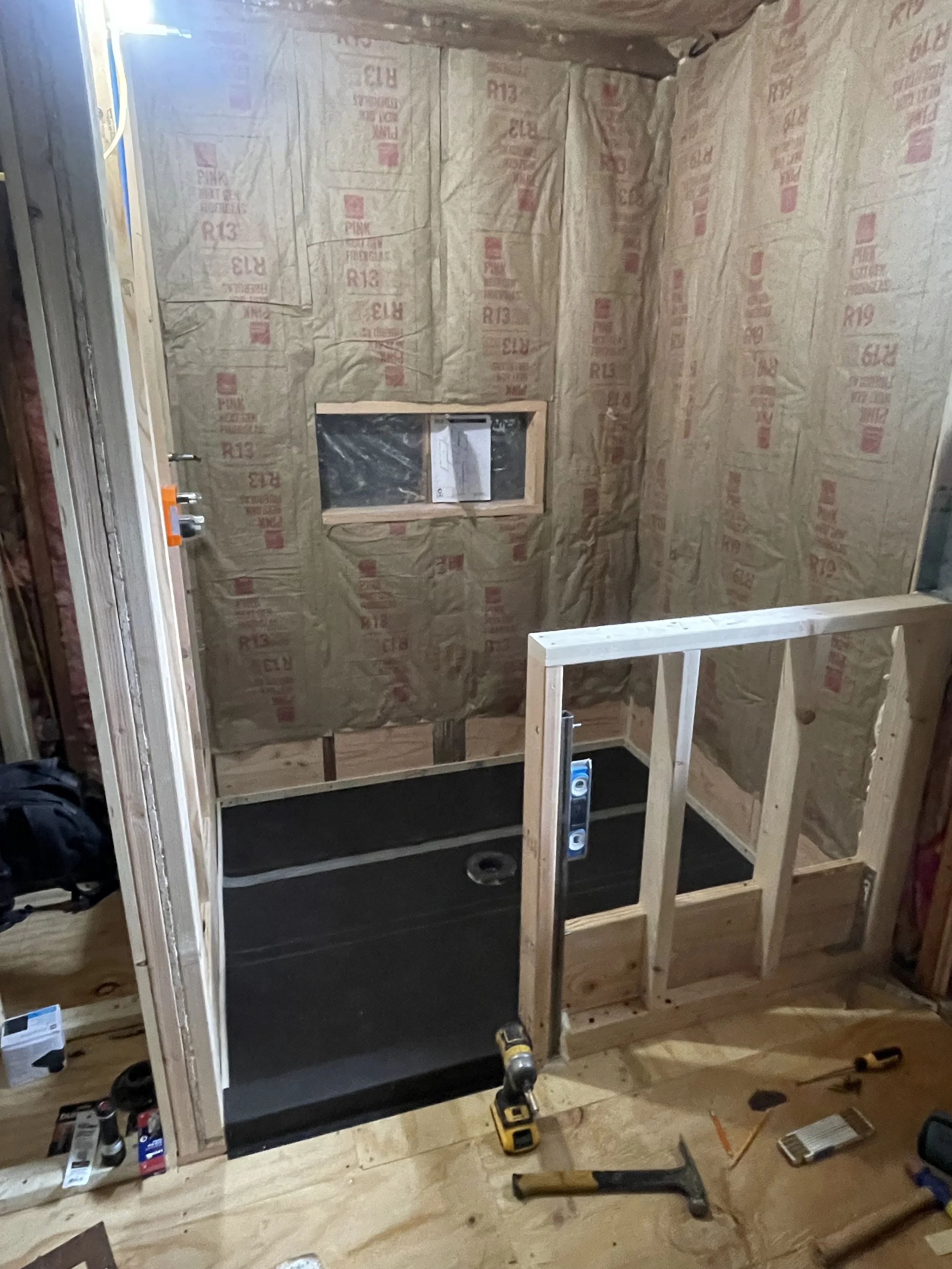 Bathroom under construction with framing, insulation, and a shower base installed.