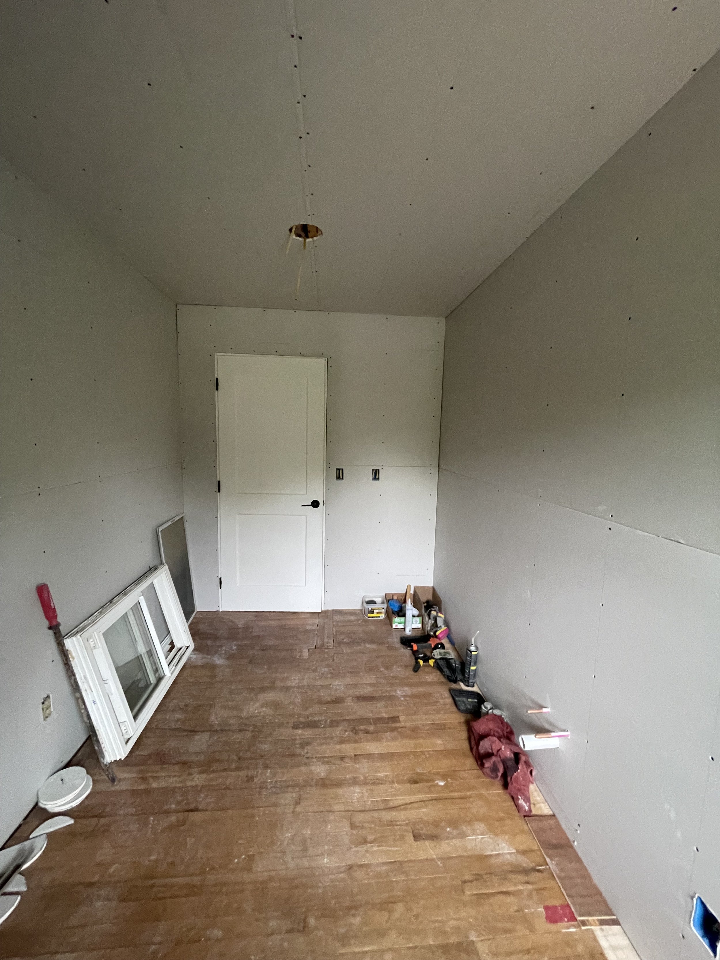 Room under construction with drywall on walls and ceiling, hardwood floor, window frame leaning against wall, and various tools on the floor.