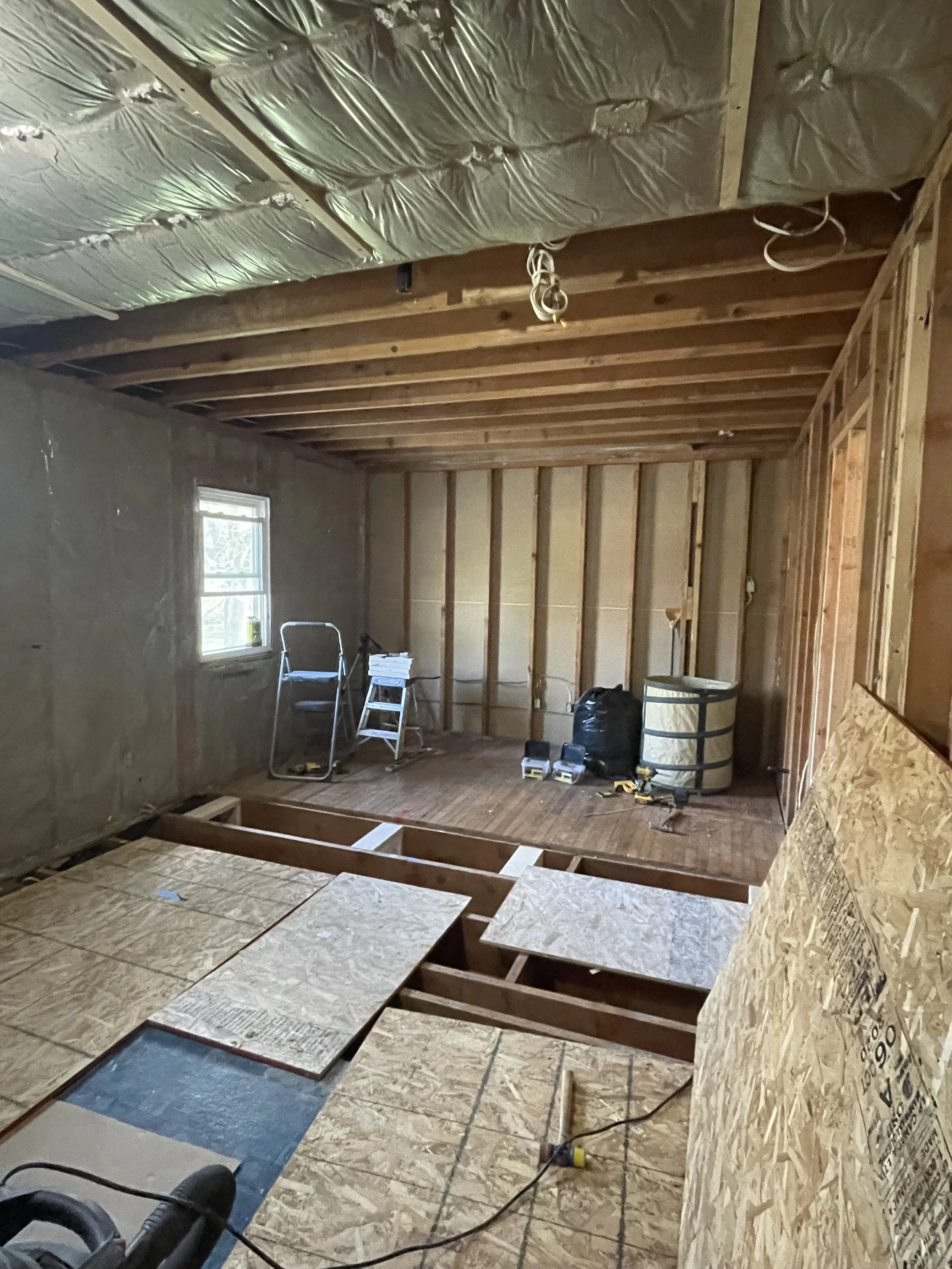 Interior of a room under construction, with exposed wooden framing, plywood floor, and insulation on the ceiling. Construction tools and materials are scattered around, including a ladder, black bag, and power tools. A small window is on the left wal