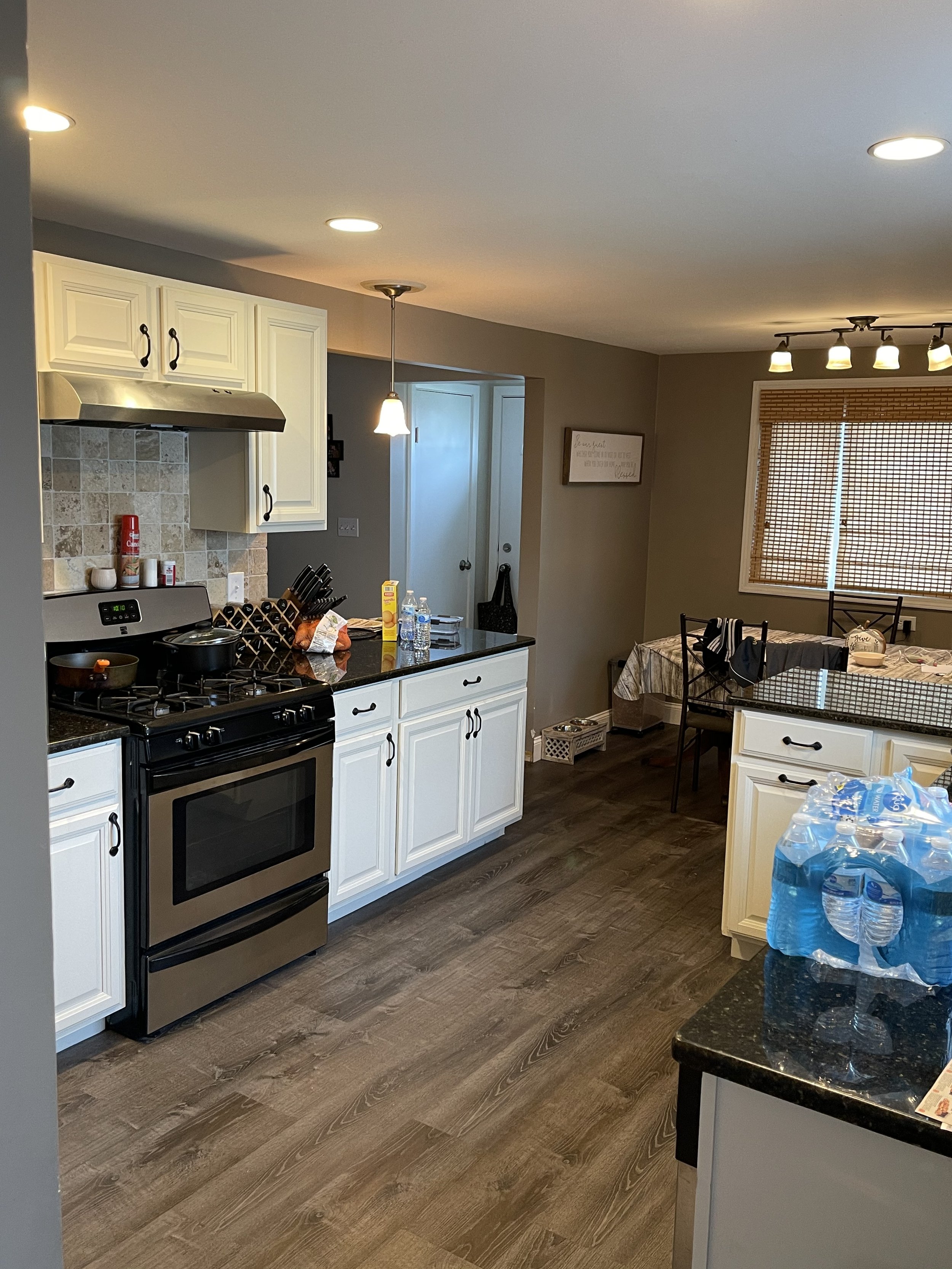 Kitchen with white cabinets, black countertops, stainless steel stove, and beige tile backsplash. There is a hanging light fixture and multiple ceiling lights. A table with chairs is in the dining area near a window with bamboo blinds. Counter with b