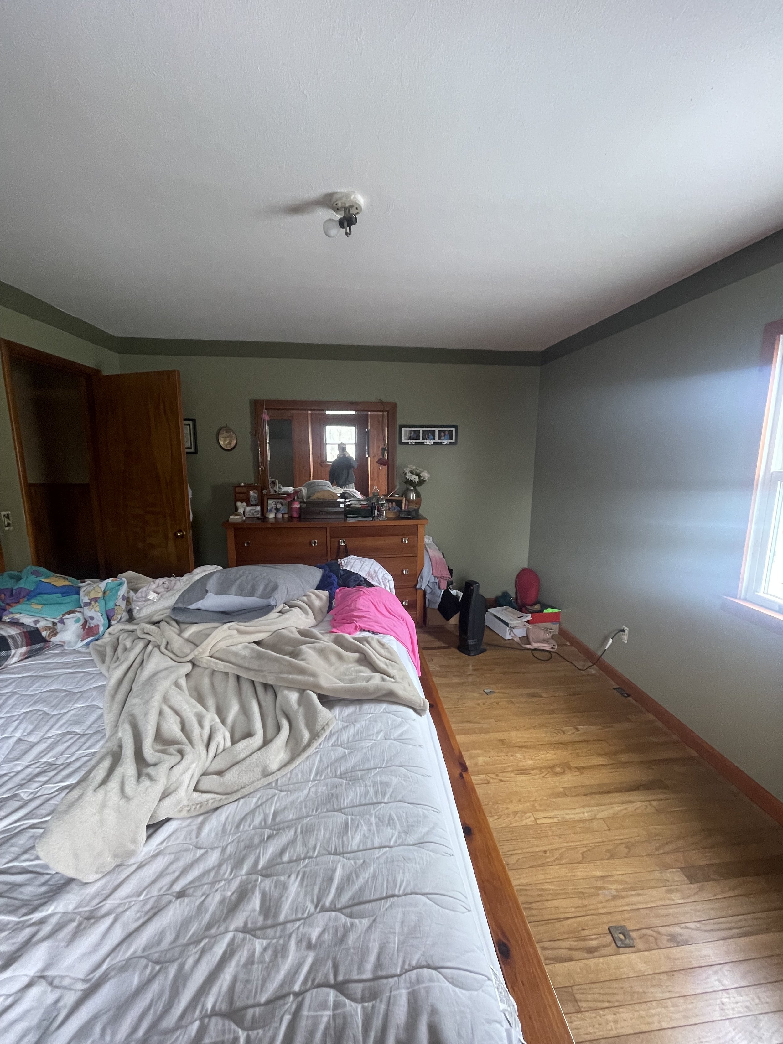 A cluttered bedroom with an unmade bed, a dresser with a mirror, and a window on the right side of the room.