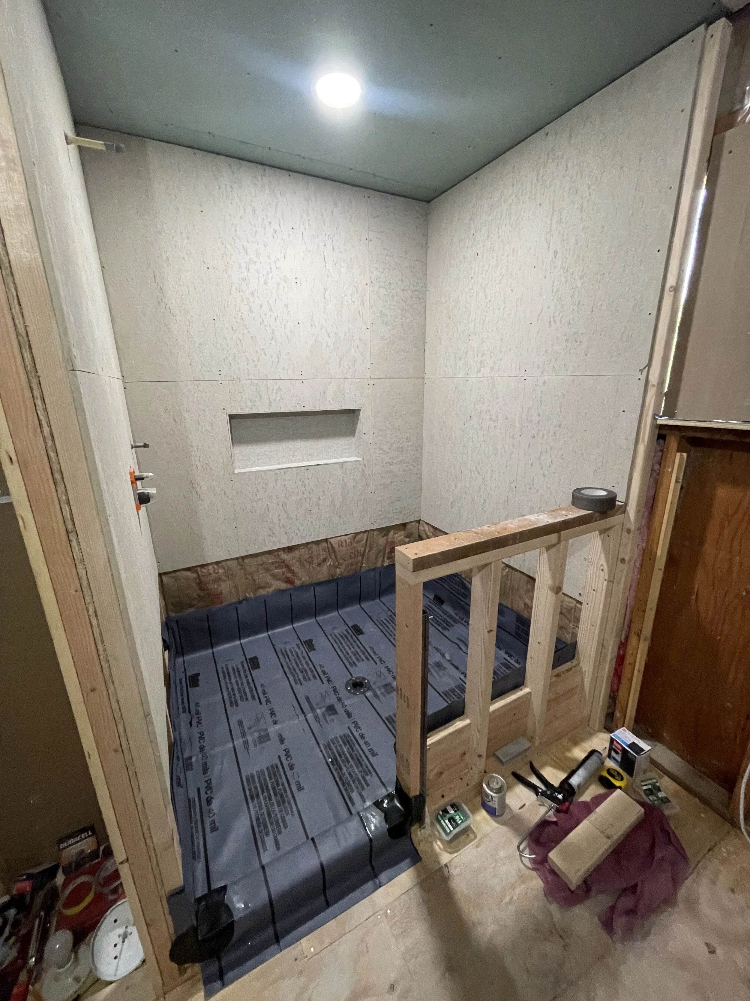 Bathroom under construction with walls installed, a recessed shelf, a partially built shower area with a drain, and tools and materials scattered around.