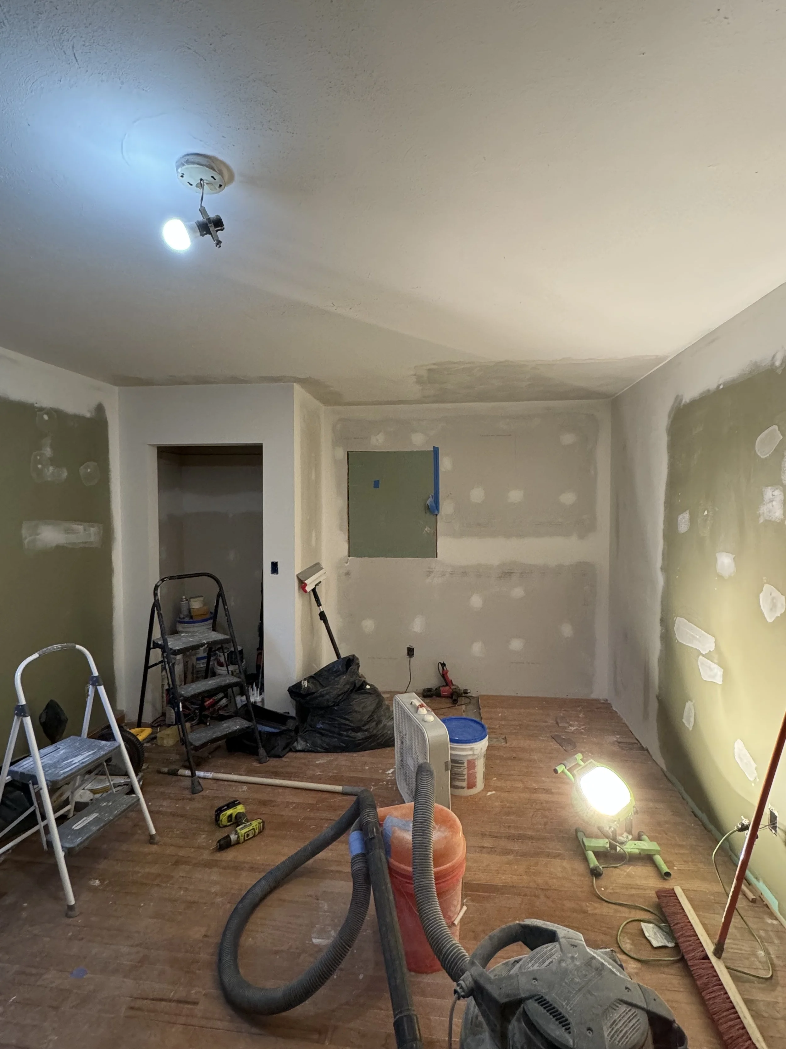 Room under renovation with construction tools, vacuum, plastic bucket, ladder, and temporary lighting.