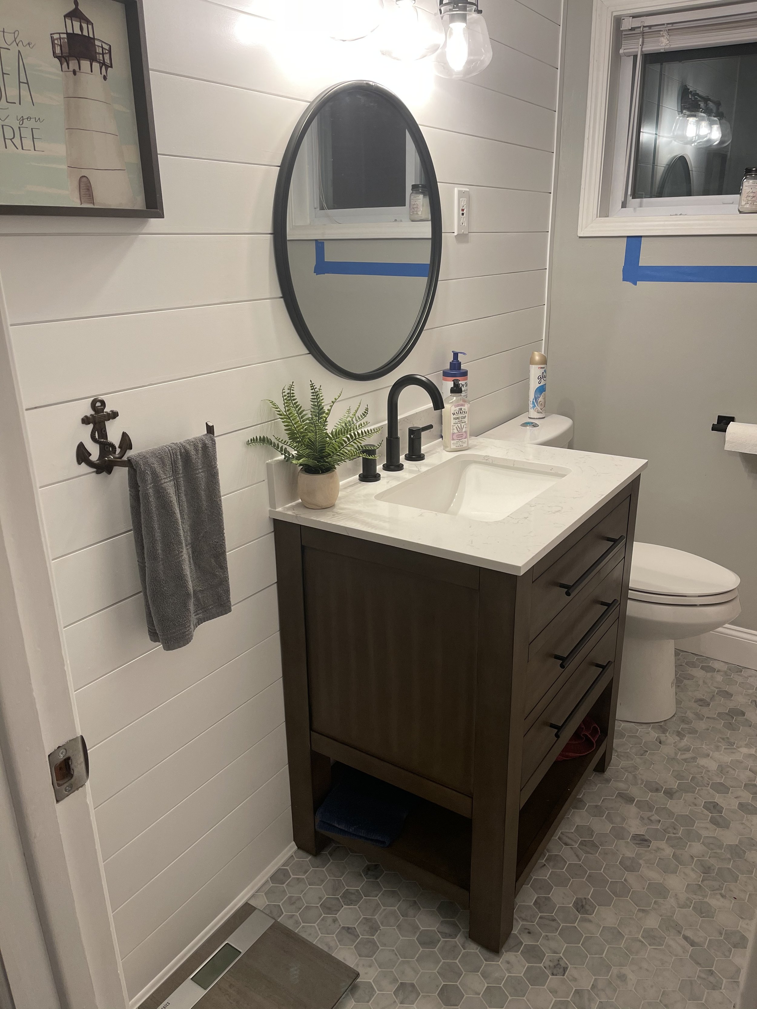 A modern bathroom with a white shiplap wall, round mirror, black faucet, and dark wooden vanity with white countertop. Small animal-themed hooks hold a towel. There are potted plants, toiletries, and a window with a blue tape marking.