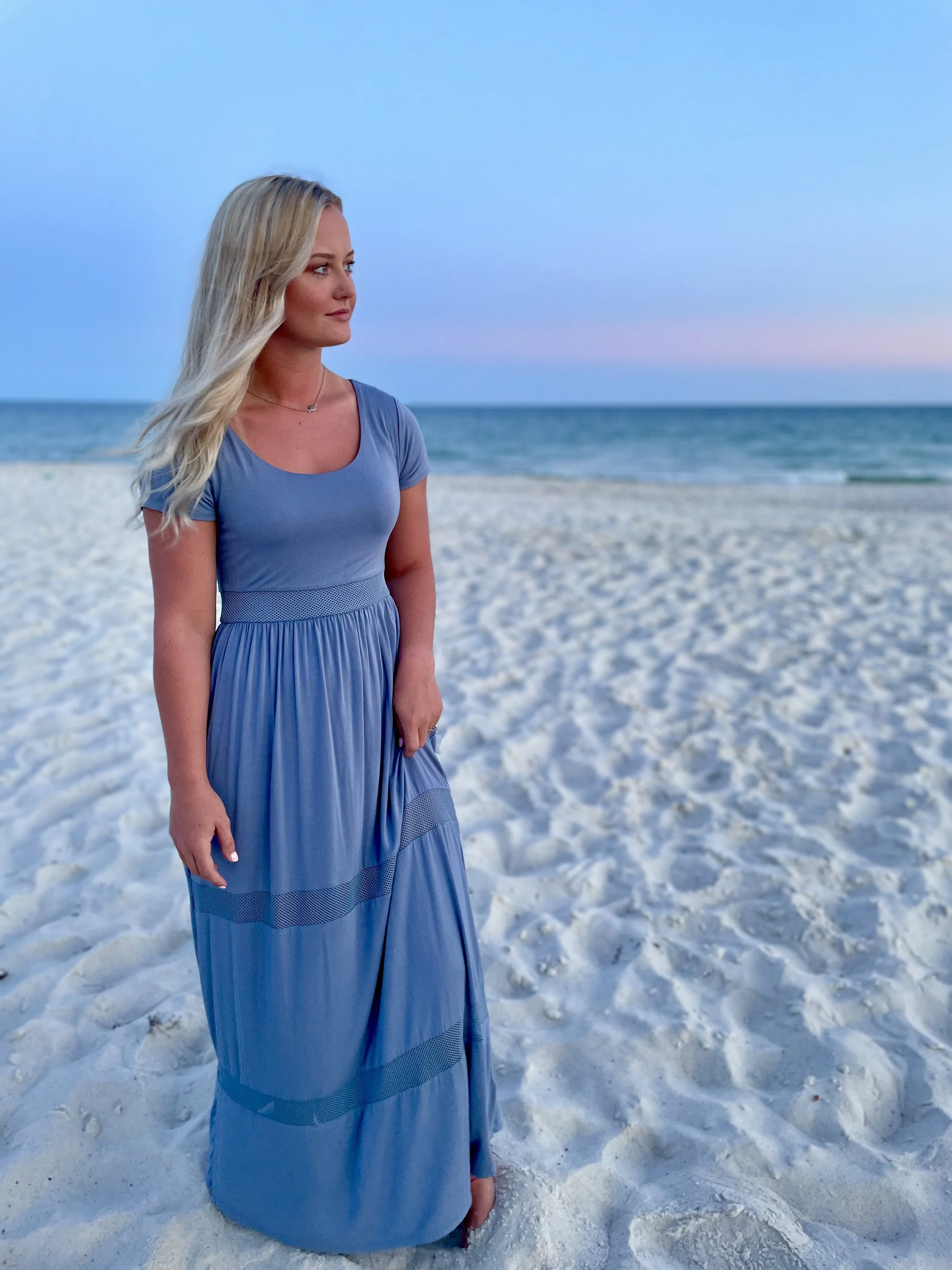 Woman in a blue dress standing barefoot on the sandy beach during sunset or sunrise, facing left with her blonde hair flowing.