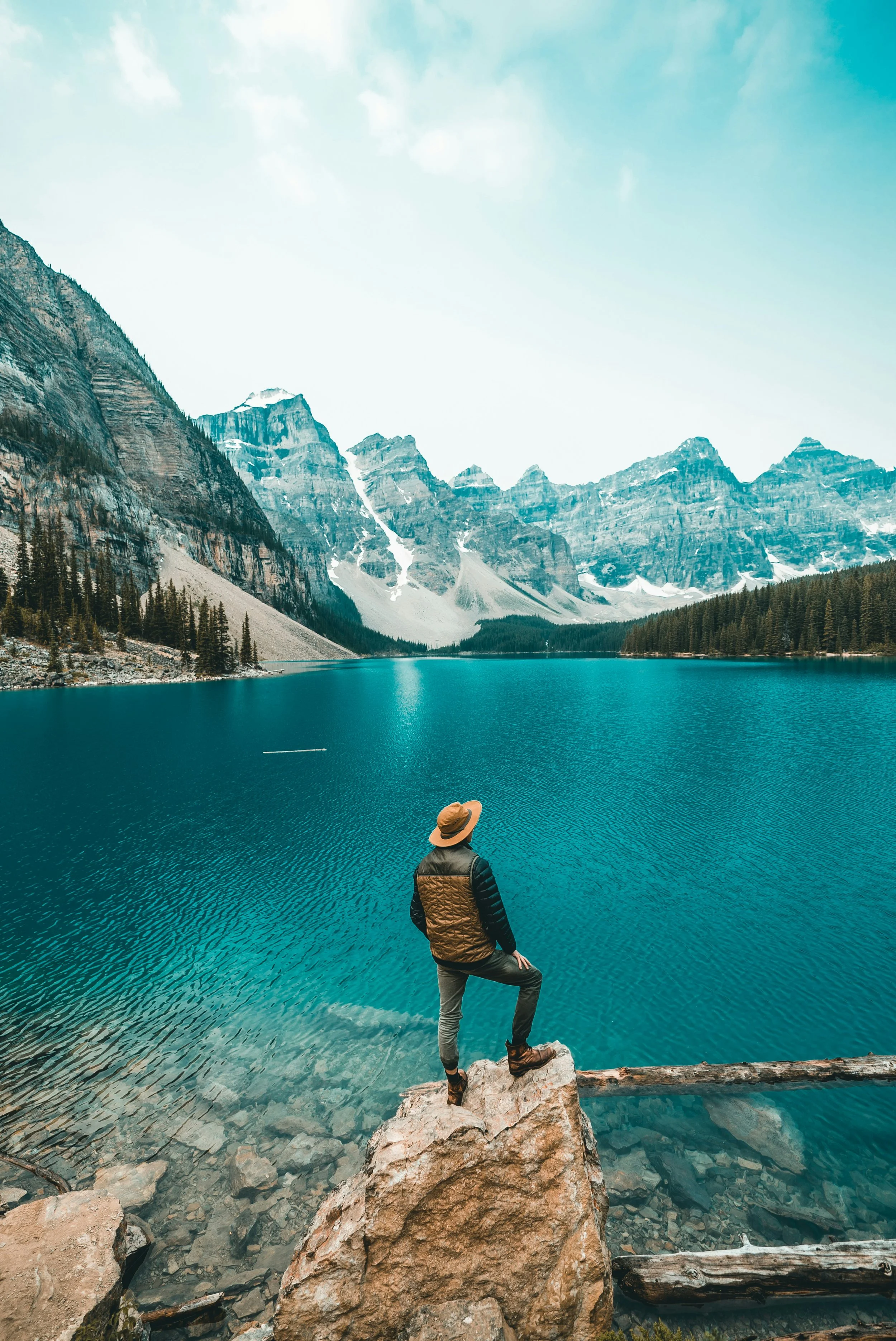 A person wearing a tan hat, black jacket, and gray pants standing on a large rock overlooking a mountain lake surrounded by pine trees and snow-capped peaks.