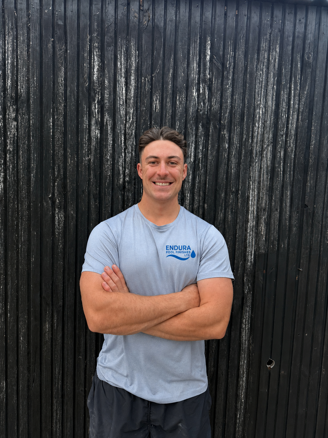 A smiling man with short dark hair, wearing a light blue t-shirt with the logo 'ENDURA POOL FINISHES LTD' on it, standing with arms crossed in front of a black wooden fence.