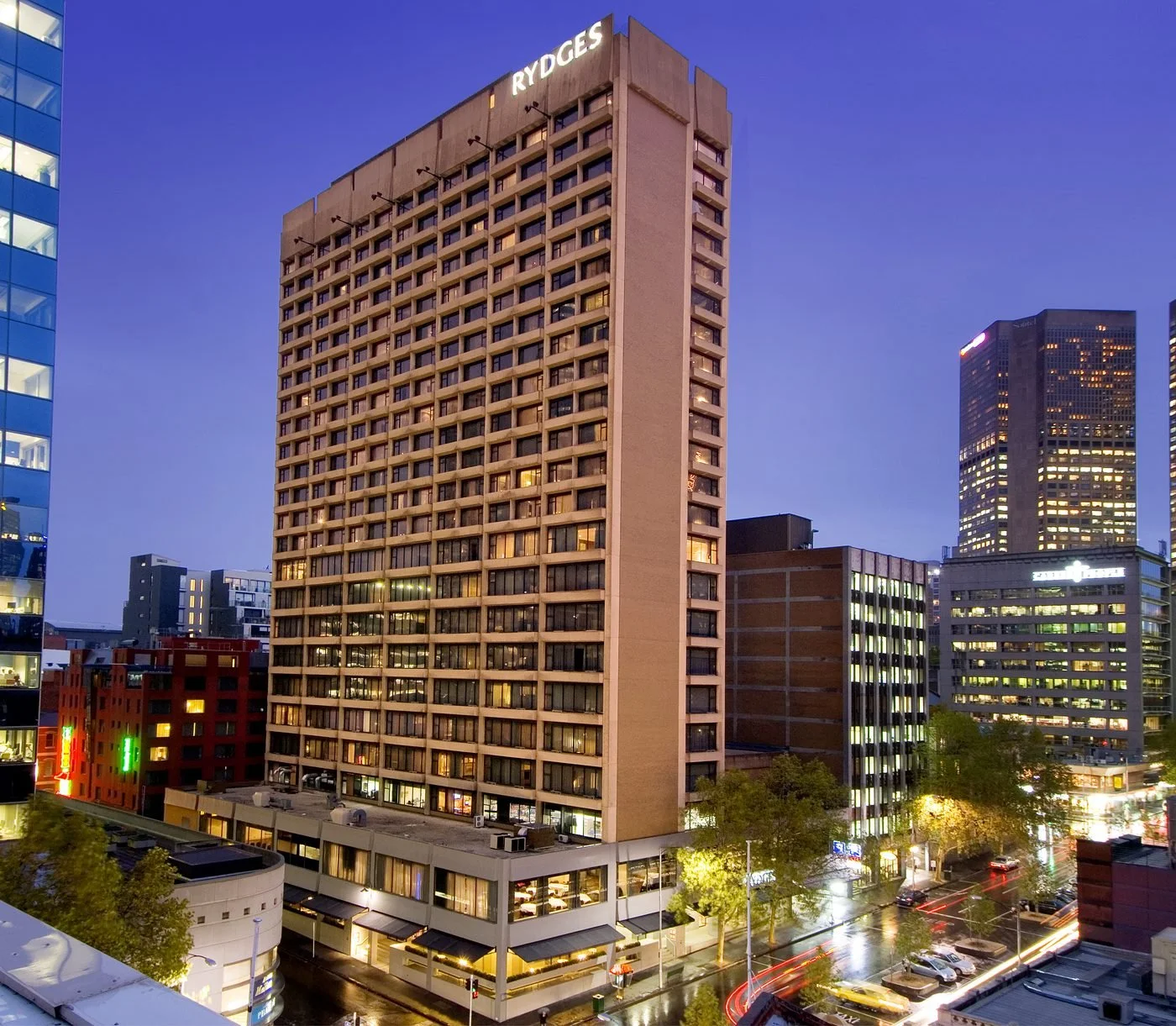 Cityscape at dusk with tall buildings, illuminated windows, and a street with cars and trees, including a prominent high-rise with RYDGES sign at the top.