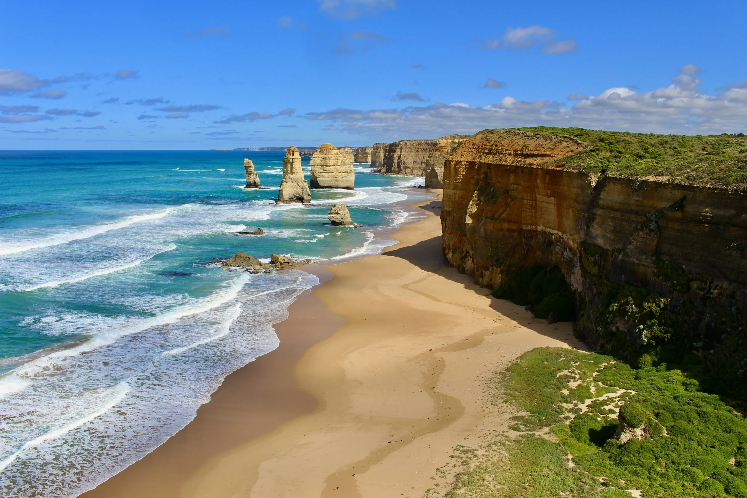 Scenic view of the Twelve Apostles, limestone stacks off the coast of the Great Ocean Road in Australia, with sandy beach, green vegetation, and blue ocean under a partly cloudy sky.