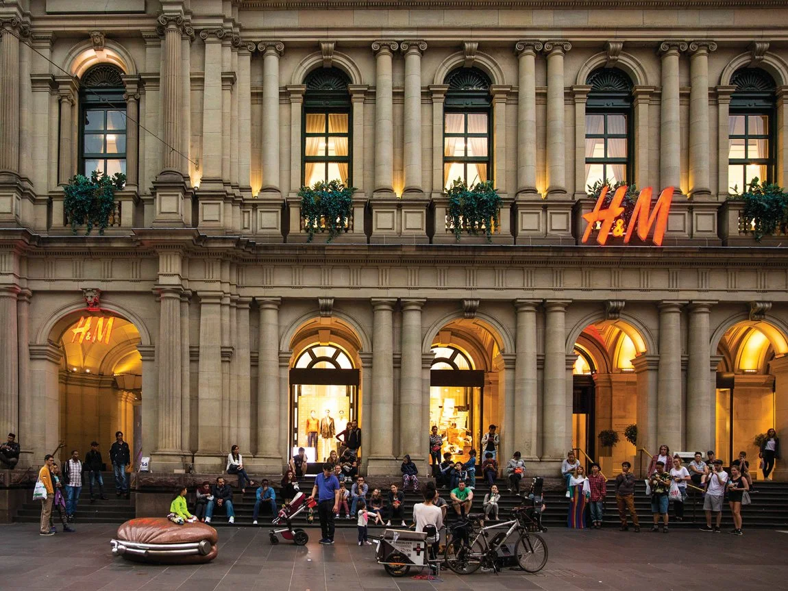 People gathered outside the H&M clothing store in a historic building with arched windows and columns, some sitting on steps and others standing, with bicycles and a seating area in front.