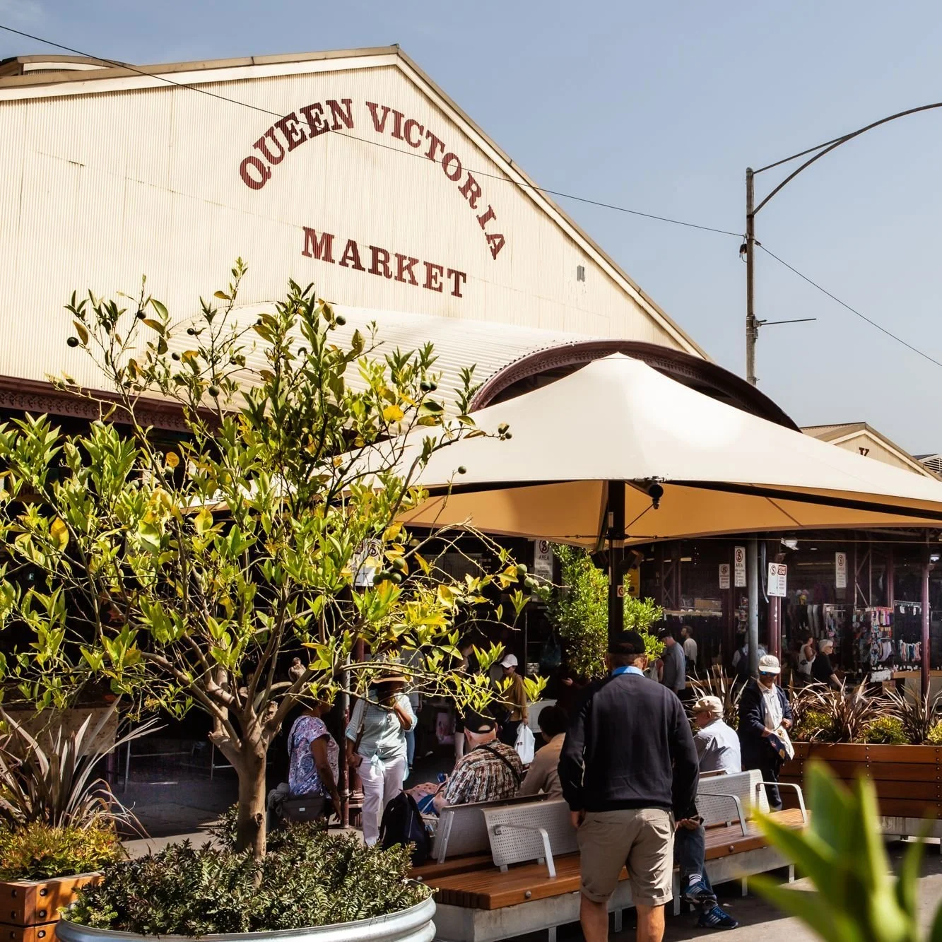 People gather outside Queen Victoria Market in Melbourne, Australia, with some sitting on benches and others walking by under large umbrellas, in front of market stalls and greenery.