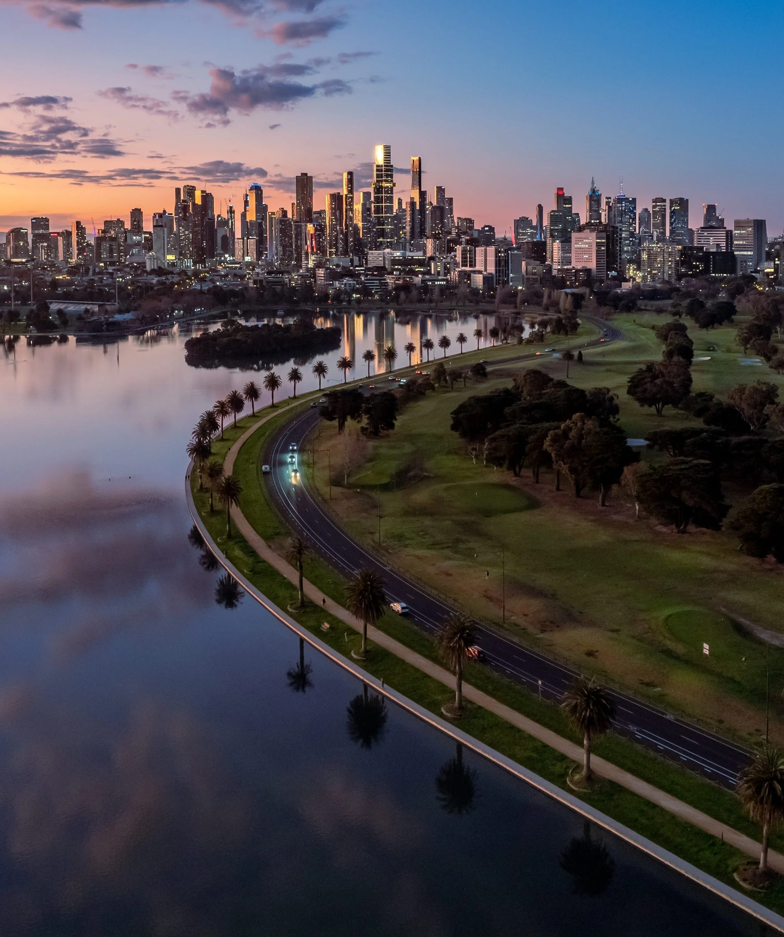 City skyline at dusk with illuminated skyscrapers, a reflecting body of water in the foreground, a curved road with palm trees, and a park area.