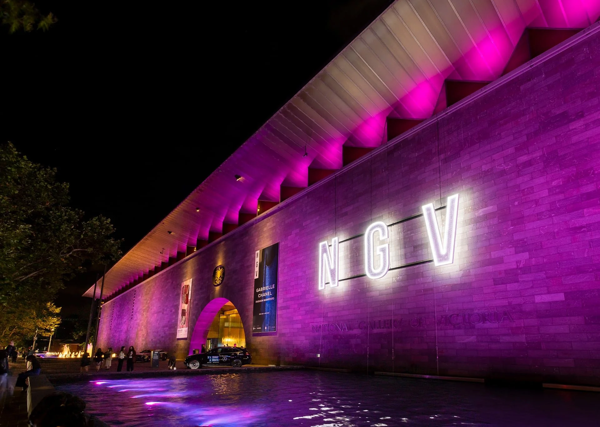Night view of the National Gallery of Victoria building illuminated with purple and pink lights, displaying large white illuminated letters 'NGV' on the wall.