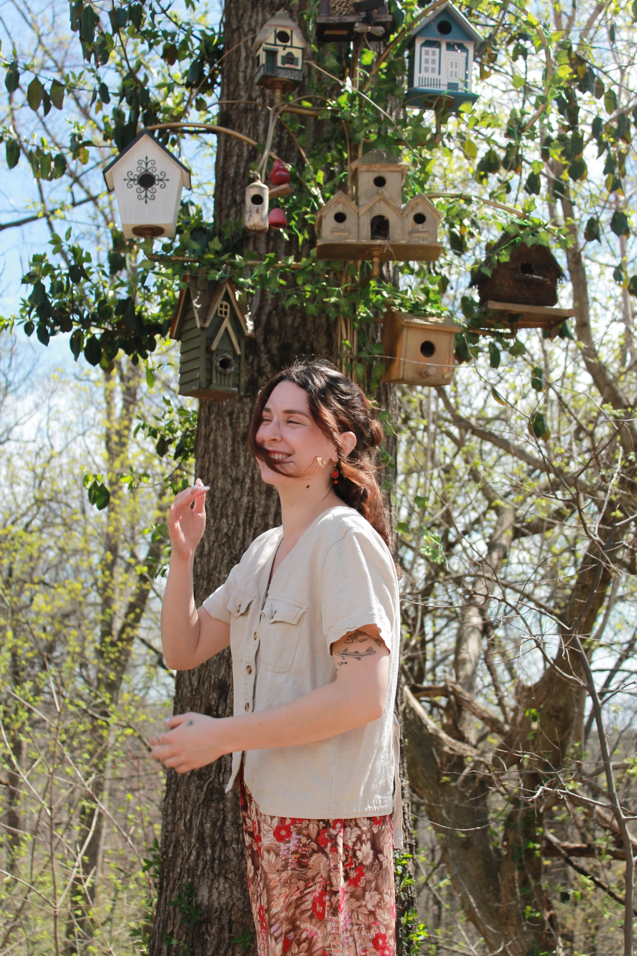 A woman standing outdoors in a wooded area with birdhouses hanging from a tree behind her, smiling and enjoying sunny weather.