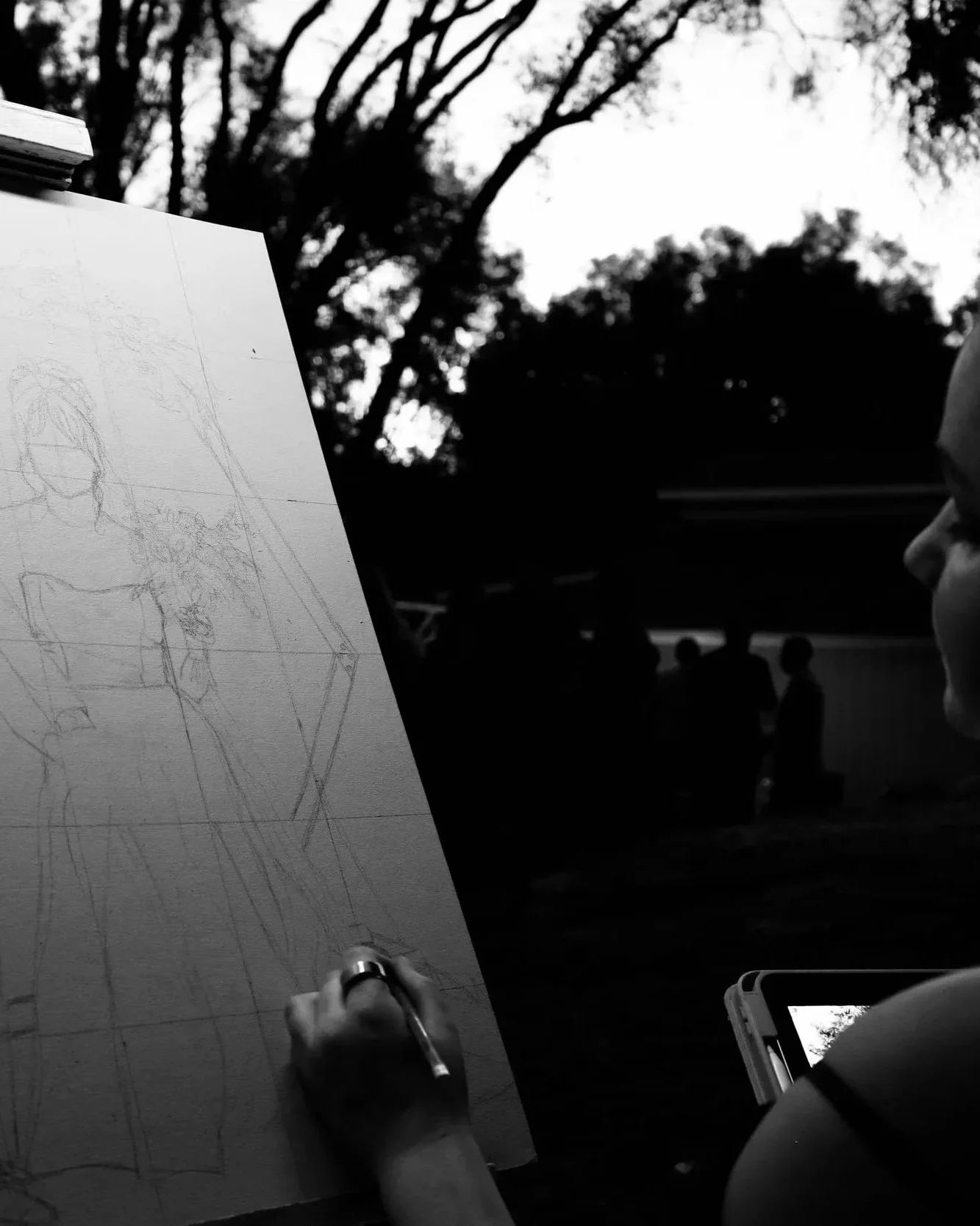 Anna Flores drawing a sketch on a large white paper with a pen, outdoors with trees and people in the background.