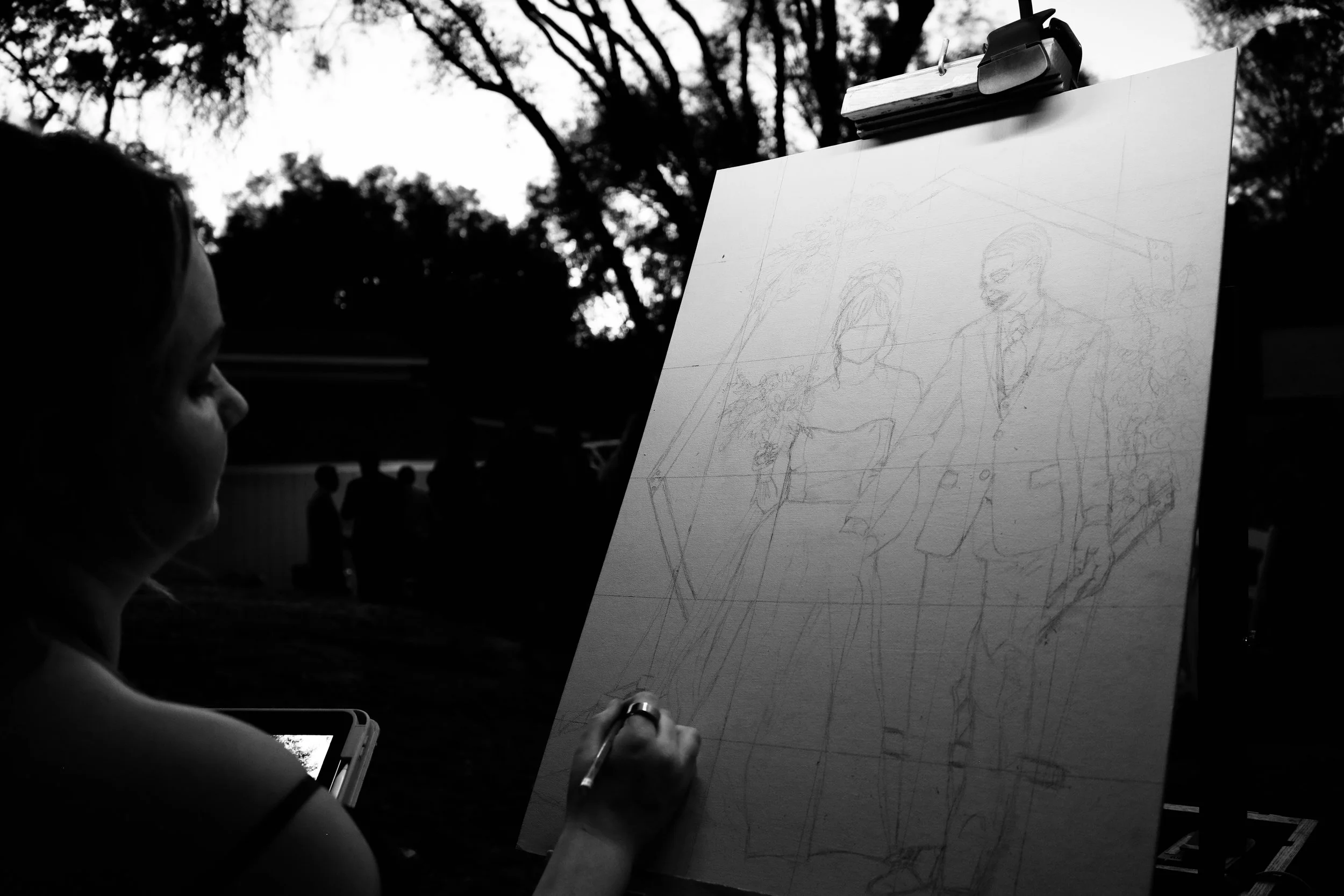 Anna Flores sketching a detailed drawing on canvas while outdoors during the day, with trees and silhouettes of people in the background.