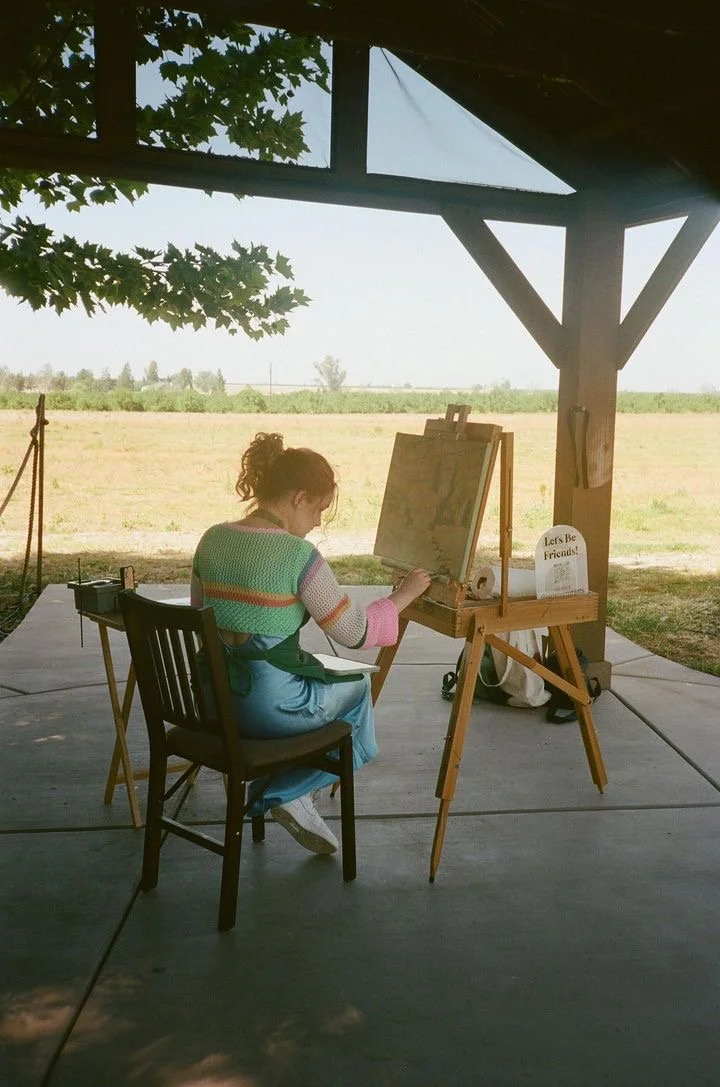 A woman painting outdoors under a wooden shelter with a view of a field in the background.