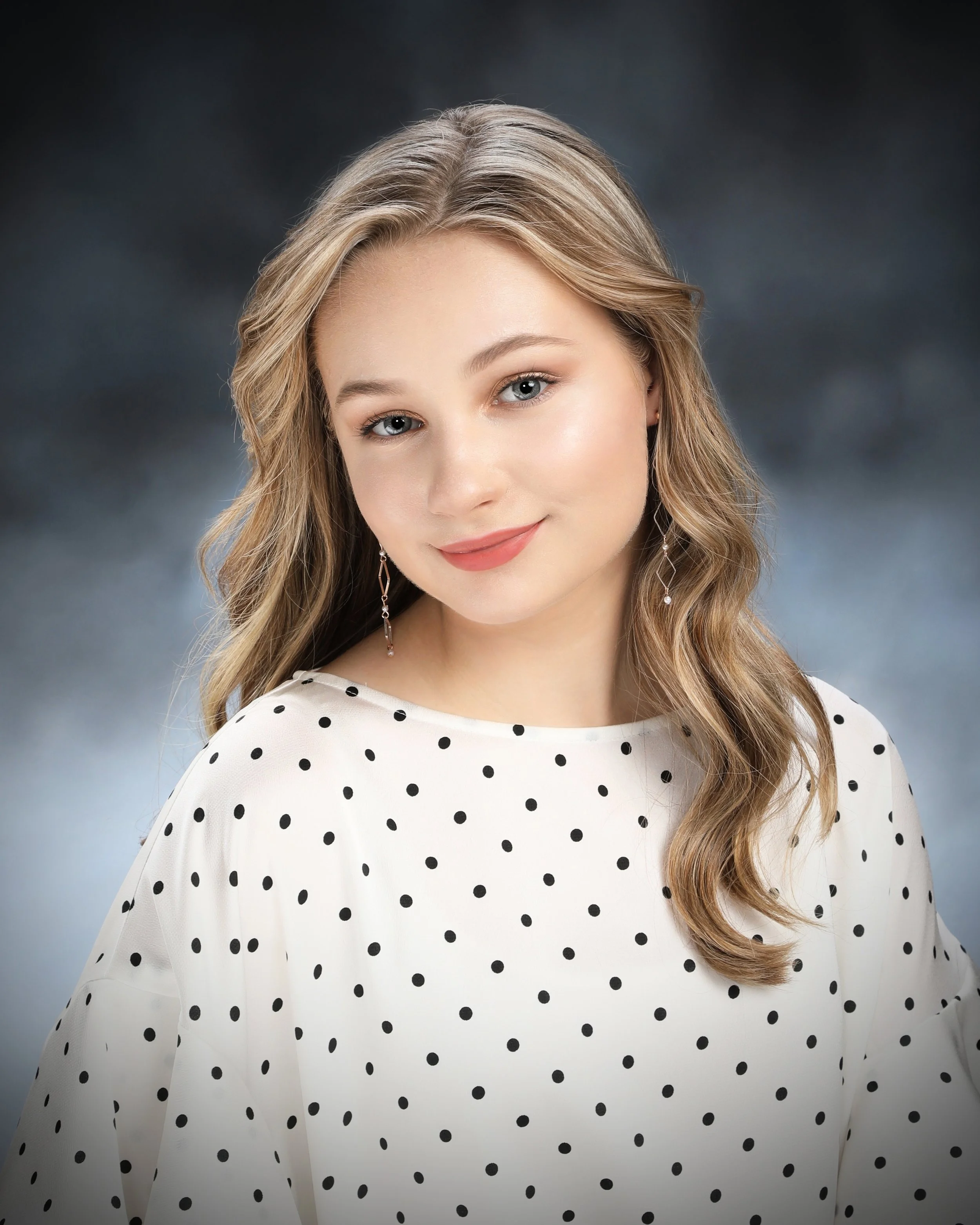 Close-up portrait of Anna Flores, a young woman with blonde wavy hair, blue eyes, wearing a white blouse with black polka dots and dangly earrings, smiling softly against a dark cloudy background.