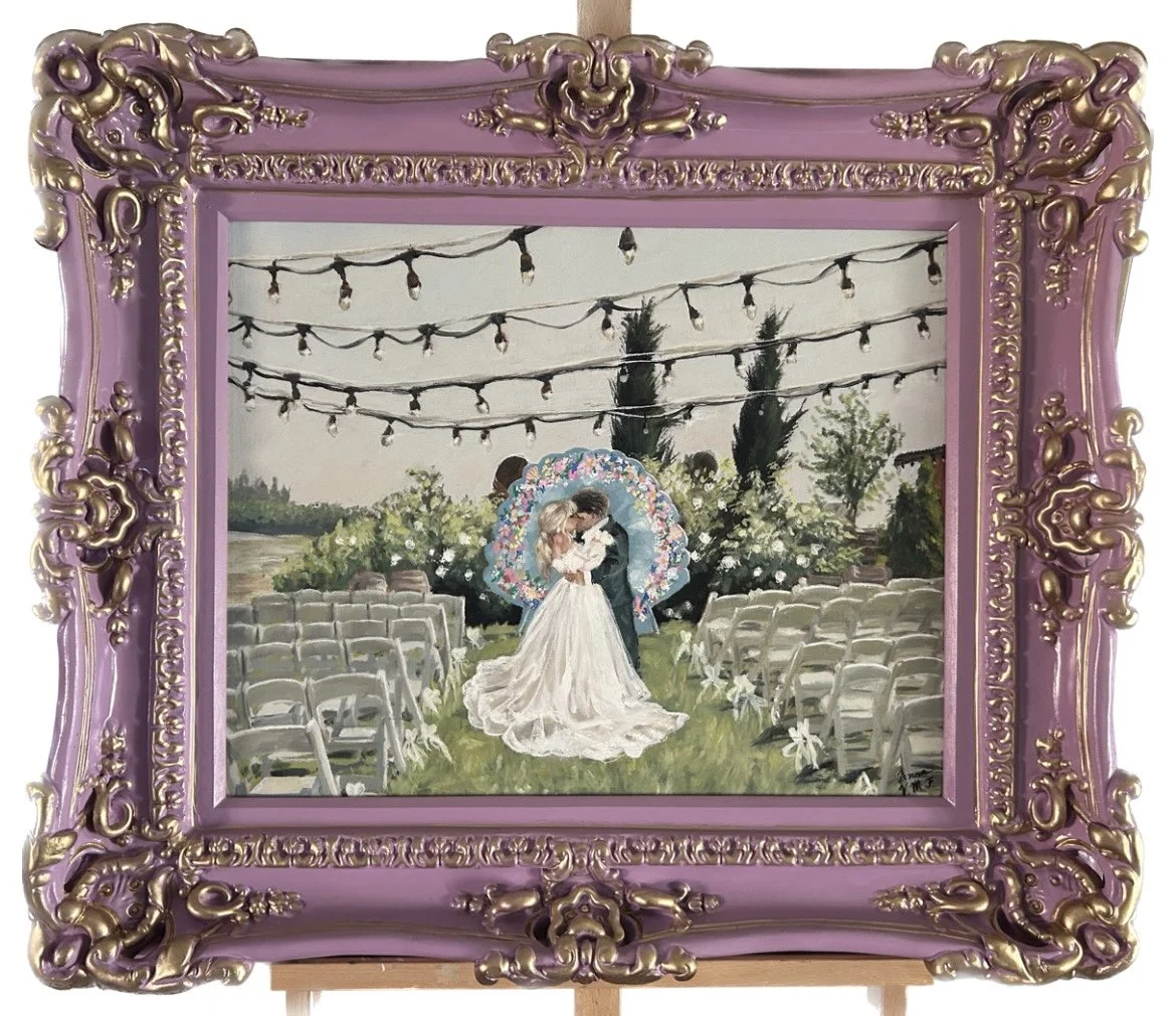 Live wedding painting of a bride and groom embracing at an outdoor wedding ceremony with string lights and trees in the background, framed in an ornate purple and gold frame.