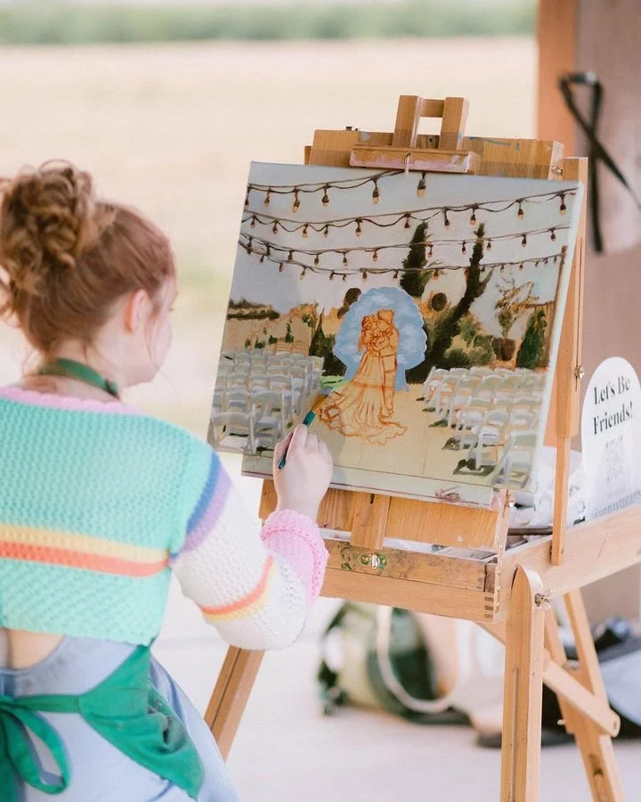 A girl painting a wedding scene on a canvas with chairs set up outdoors, string lights overhead, and a scenic background.