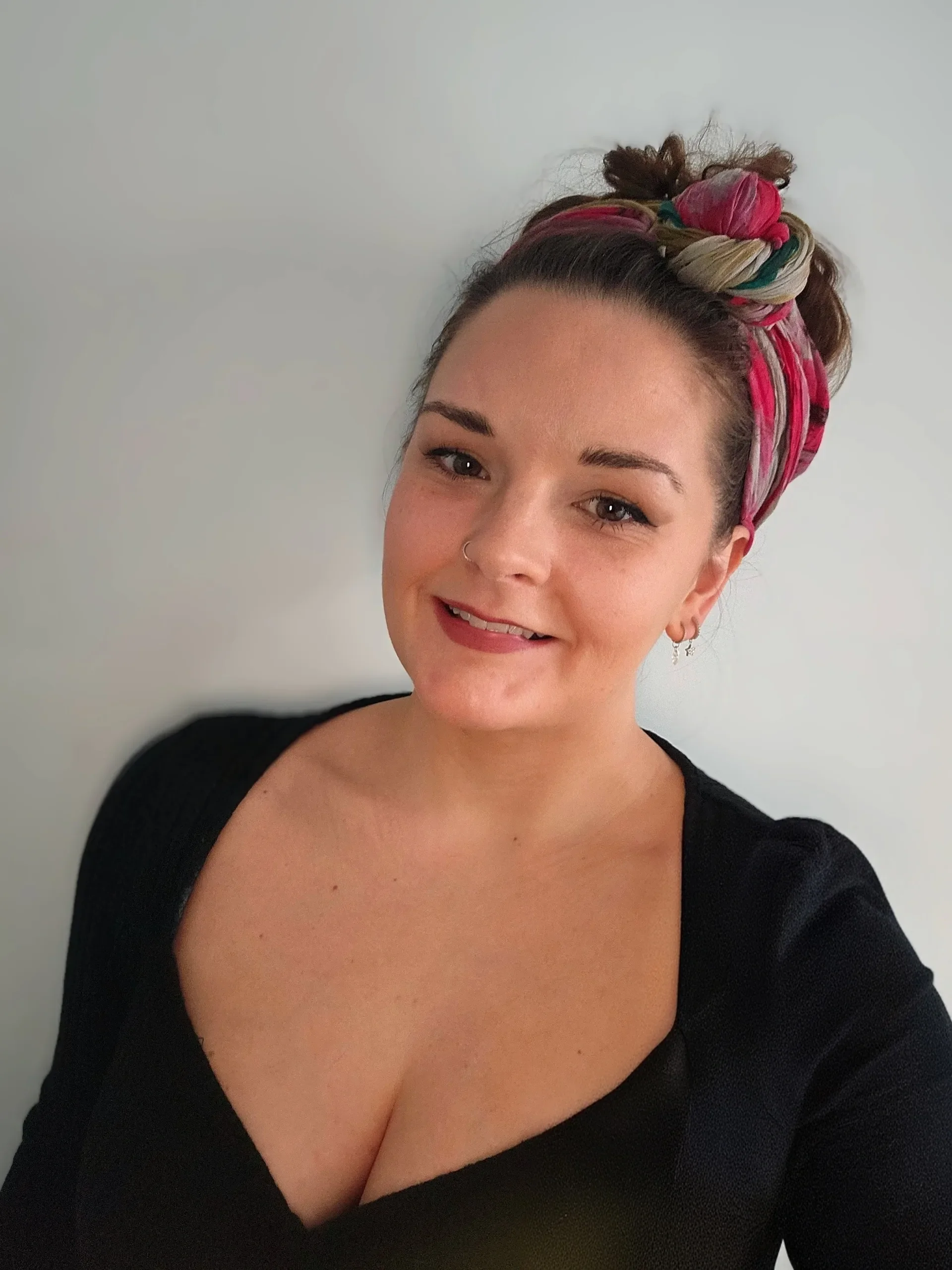A woman smiling with dark hair tied up in a bun wrapped with a colorful headscarf, wearing a black top with a deep neckline, standing against a plain white wall.