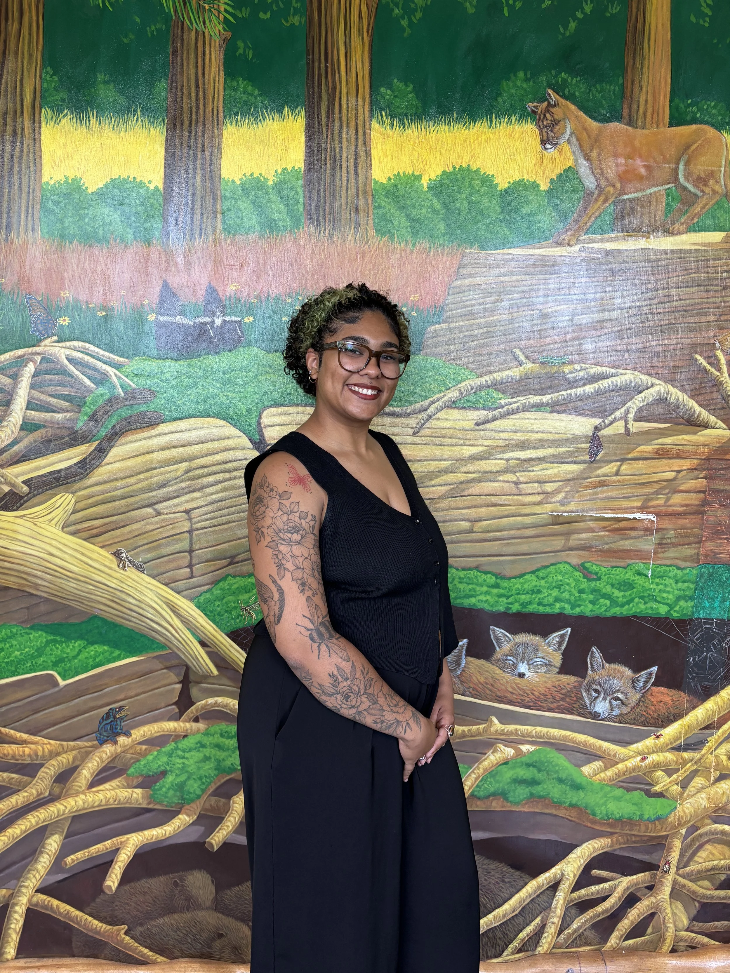 A woman with tattoos on her left arm, wearing glasses and a black sleeveless top, smiling in front of a colorful mural depicting a forest scene with wolves, a mountain lion, trees, and other wildlife.