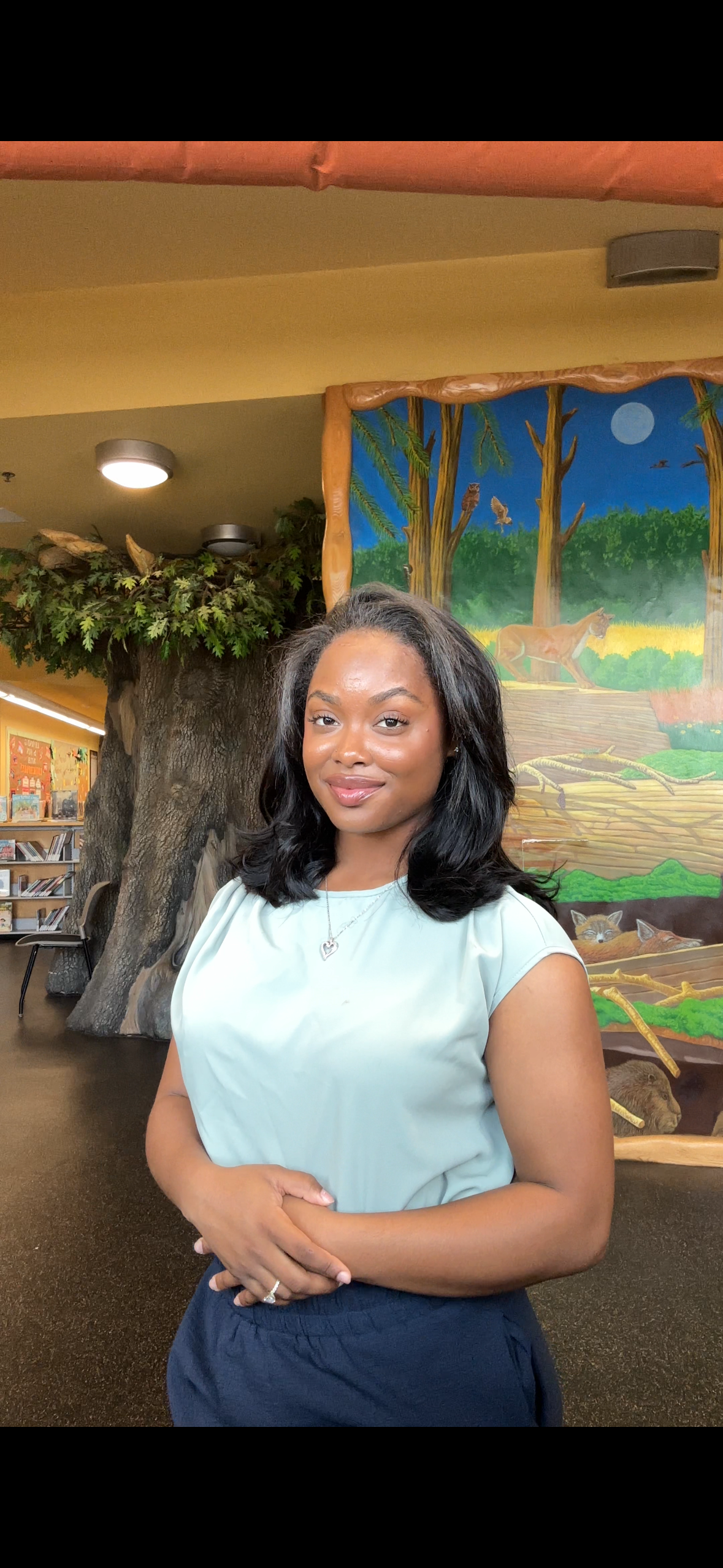 Young woman with black hair and a light blue top standing indoors with a colorful jungle mural and large tree prop in background.