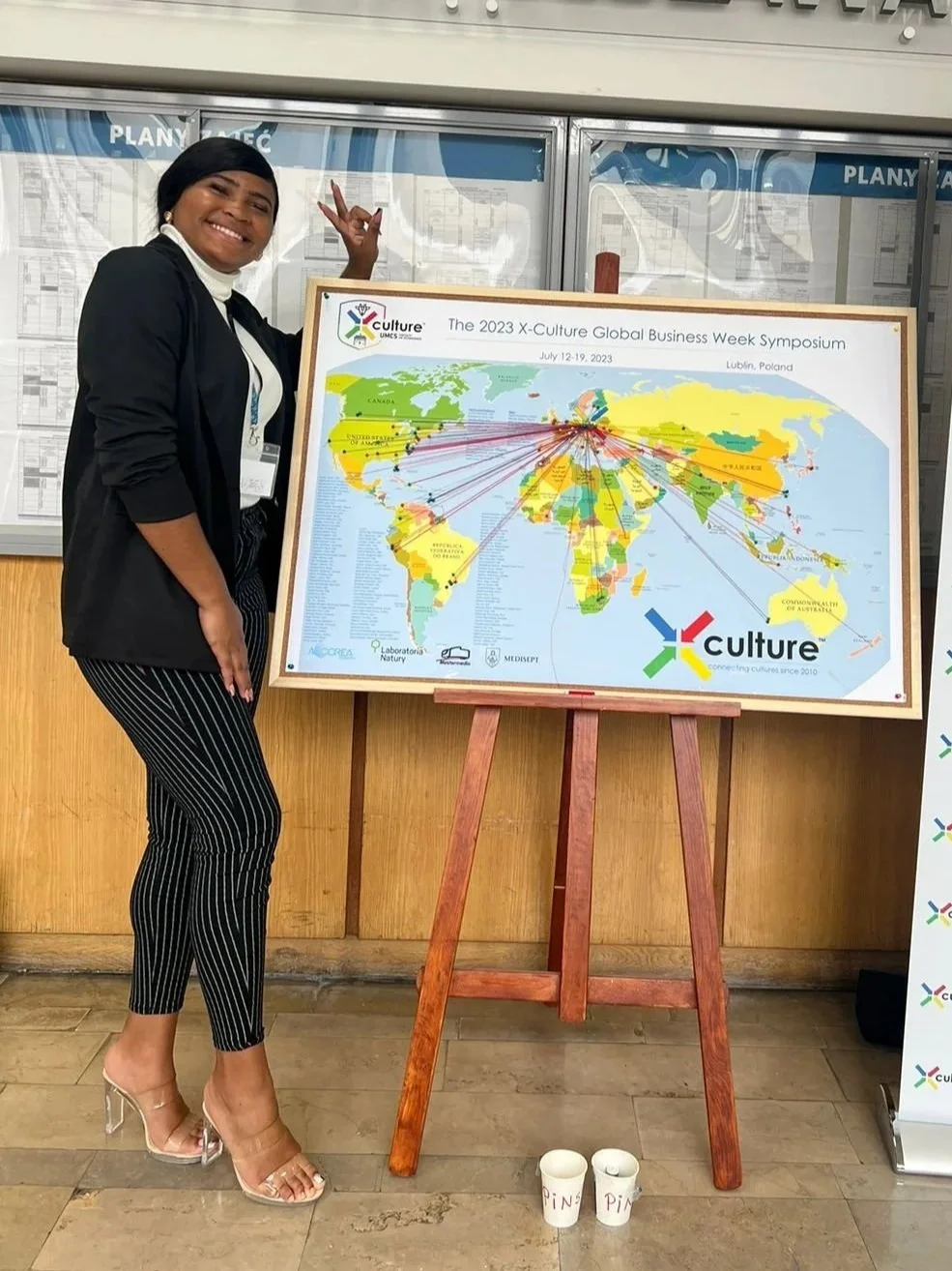A woman smiling and making a peace sign next to a world map on an easel at the 2023 X-Culture Global Business Week Symposiumm, held in Poland from July 12-19, 2023.