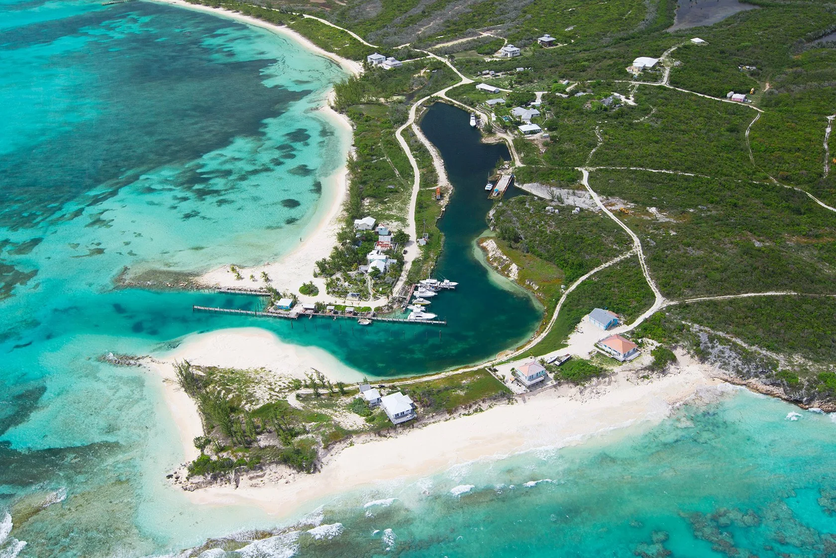 Aerial view of a coastal area with turquoise waters, white sandy beaches, houses, a marina with boats, and lush green hills with winding pathways.