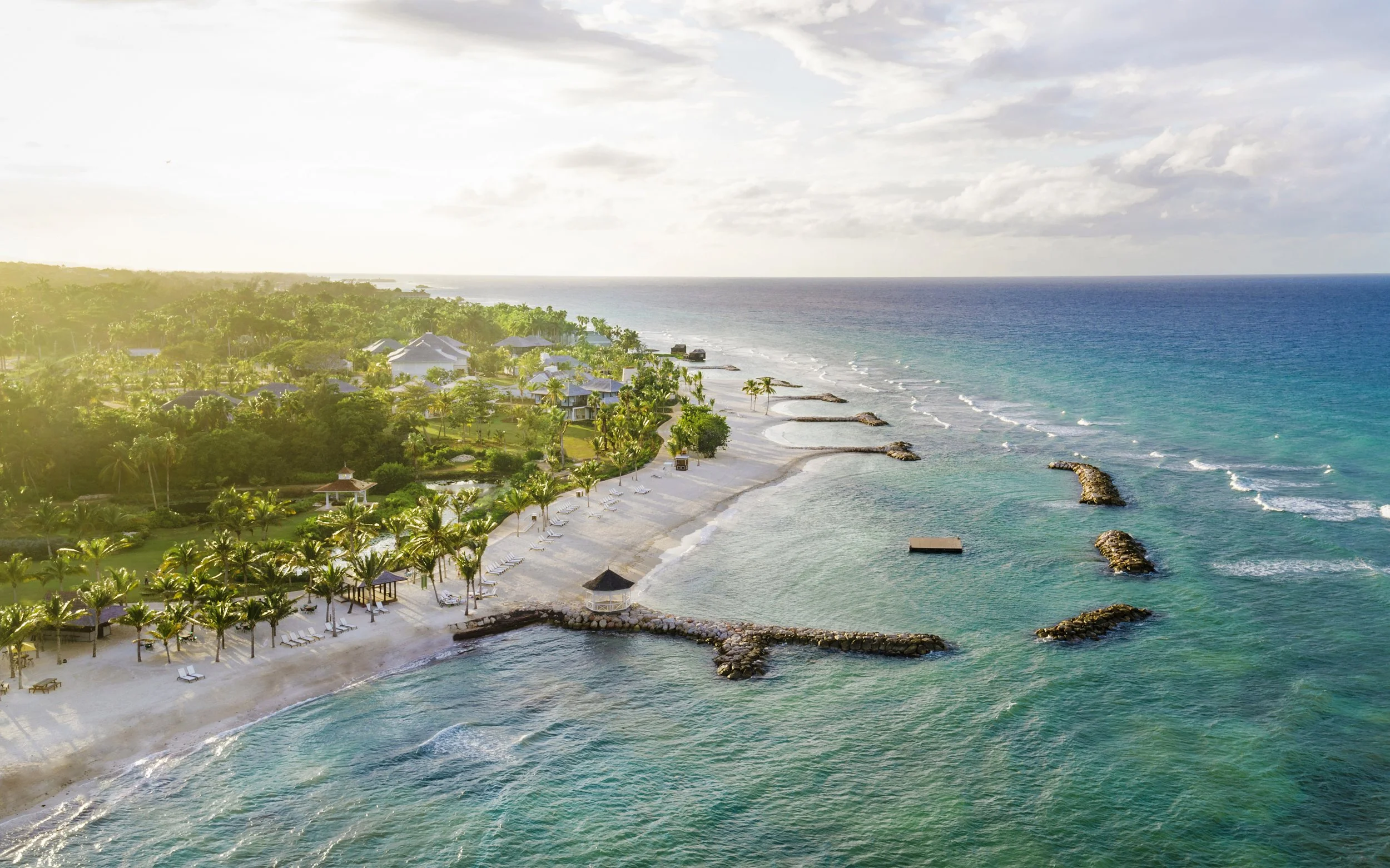 Aerial view of a tropical beach resort with white sandy beaches, palm trees, lounge chairs, small huts, and stone breakwaters extending into the ocean, under a partly cloudy sky.