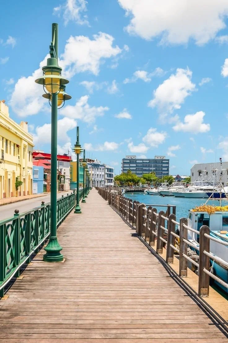 Wooden pier with green lampposts, adjacent to colorful buildings and boats docked by the water, under a partly cloudy sky.