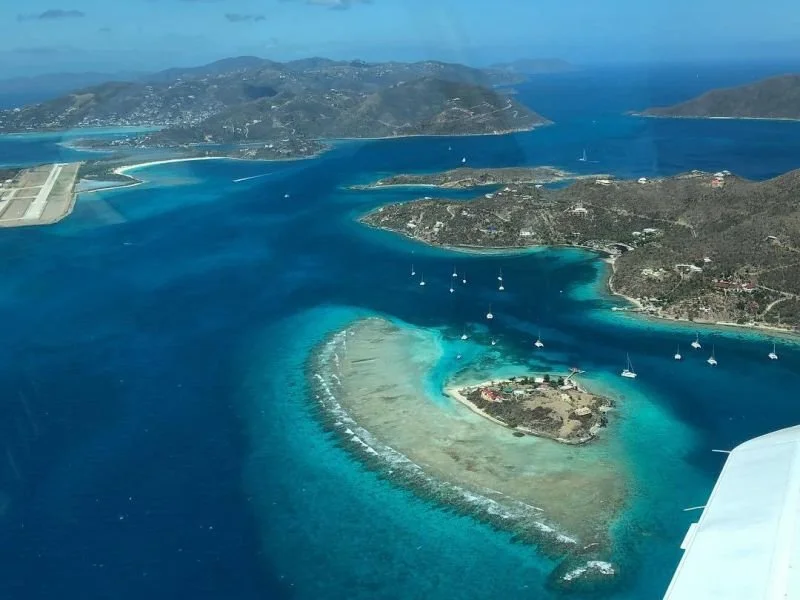 Aerial view of a coastal area with turquoise waters, small islands, sailboats, and a shoreline with hills and greenery.