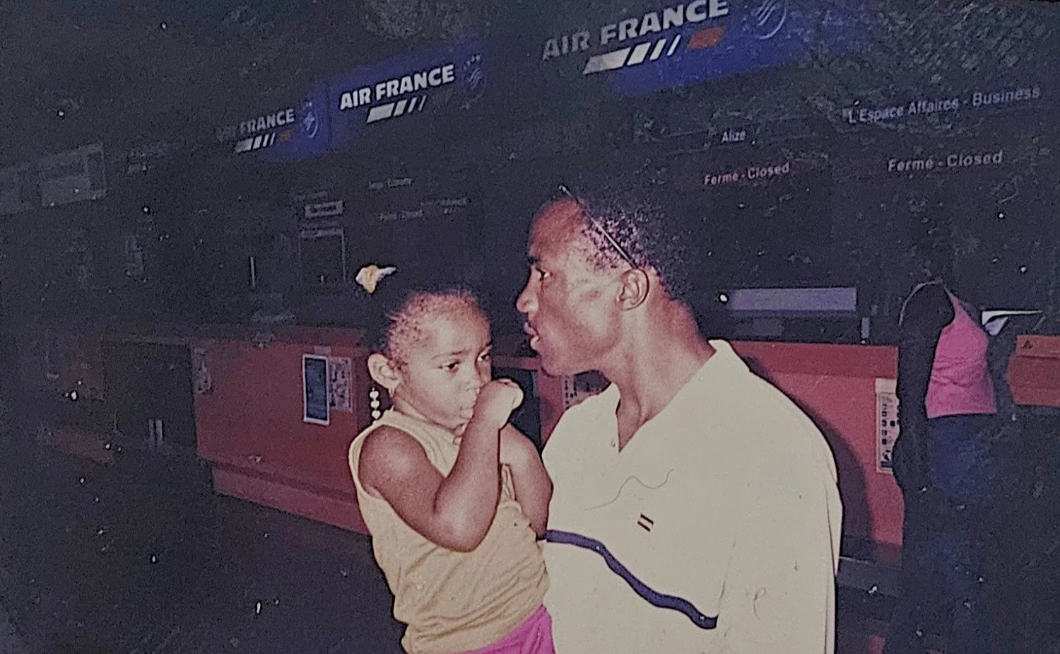 A woman holding a young girl at an airport near Air France signs. The woman is speaking to the girl, who looks thoughtful and is sucking her thumb.