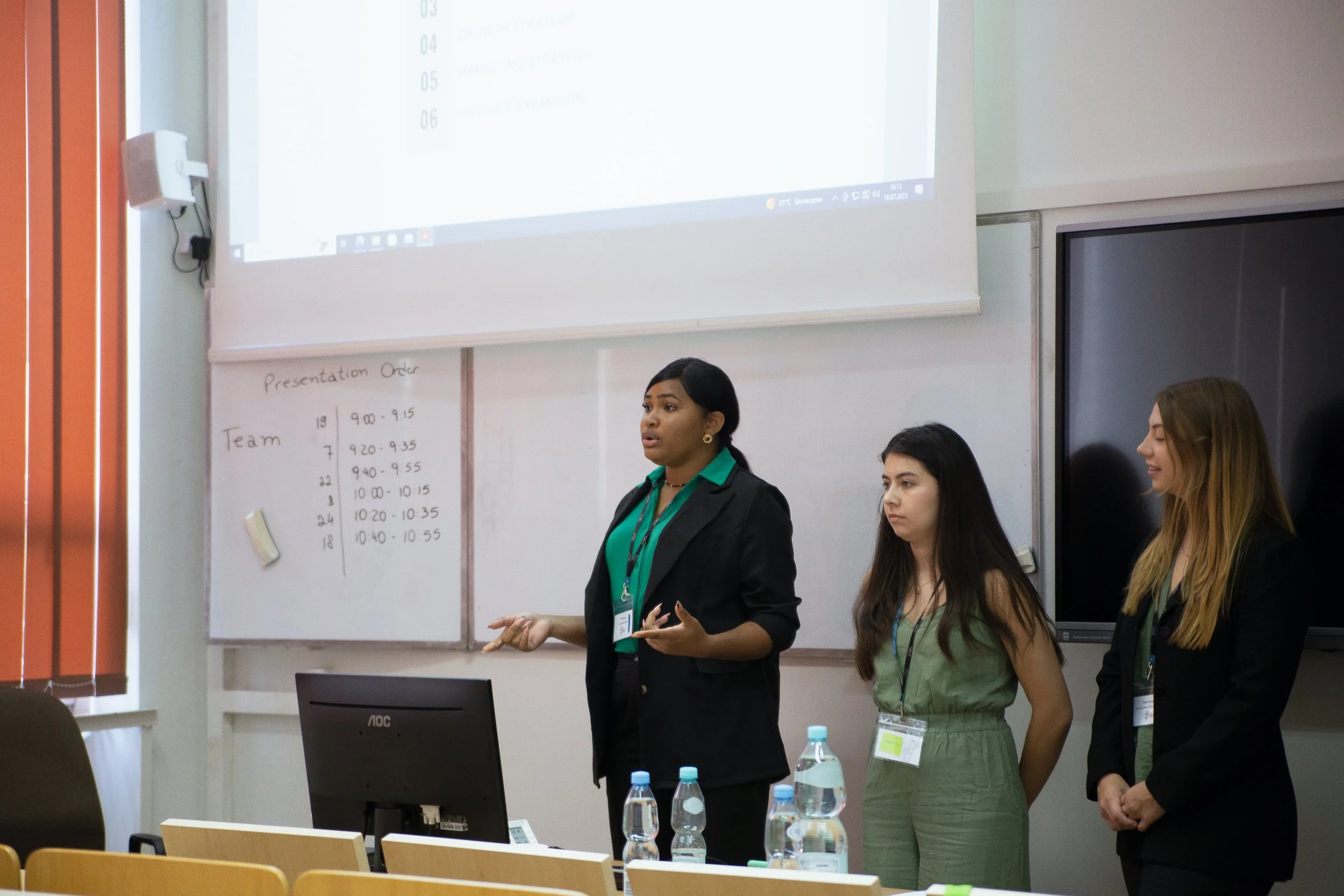 A woman in a black blazer and green shirt giving a presentation in a classroom or conference room, with three other women listening nearby. There is a whiteboard with writing on it and a large screen above displaying a list.
