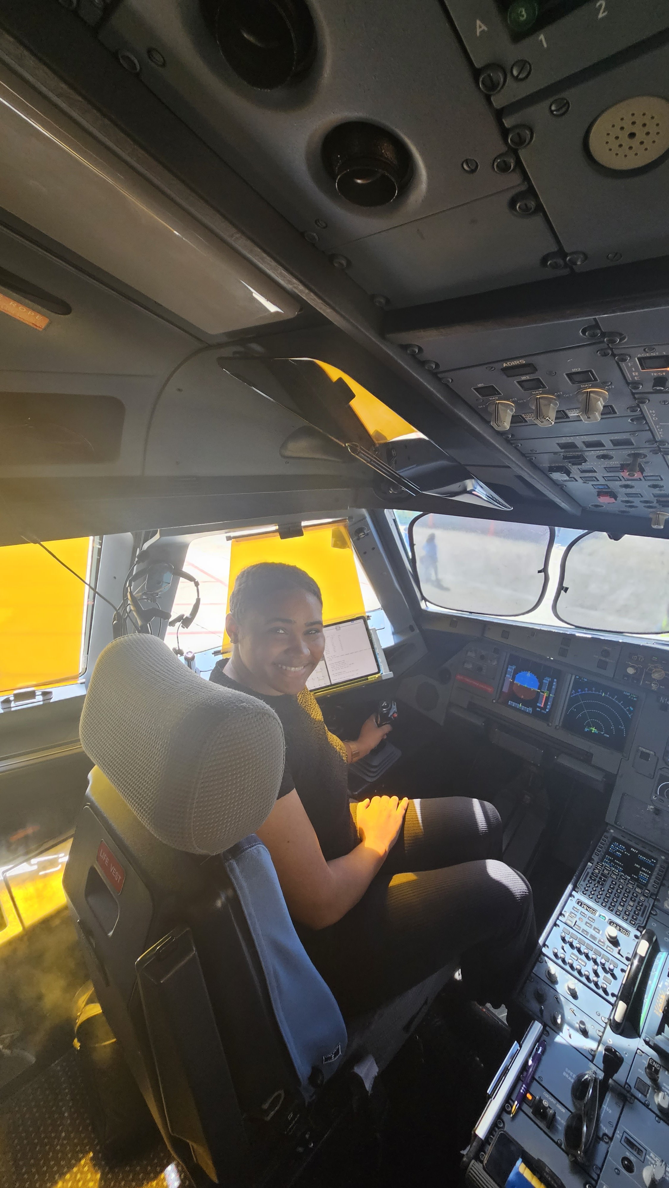 A woman sitting in the cockpit of an airplane, smiling at the camera, with the pilot's chair, cockpit controls, and aircraft windshield visible.