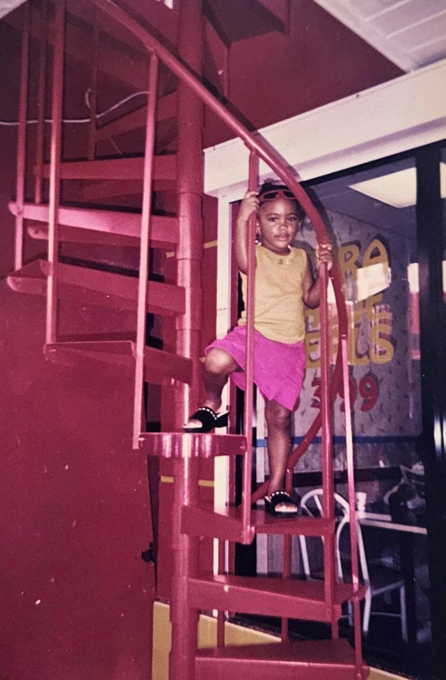 Young girl standing on a red spiral staircase inside a building, wearing a yellow top, pink skirt, and black shoes, holding onto the railing.