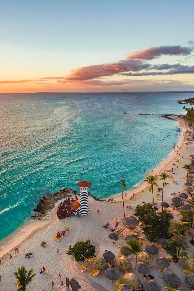 Aerial view of a tropical beach with turquoise water, a black and white striped lighthouse, palm trees, beach umbrellas, and people enjoying the sunset.