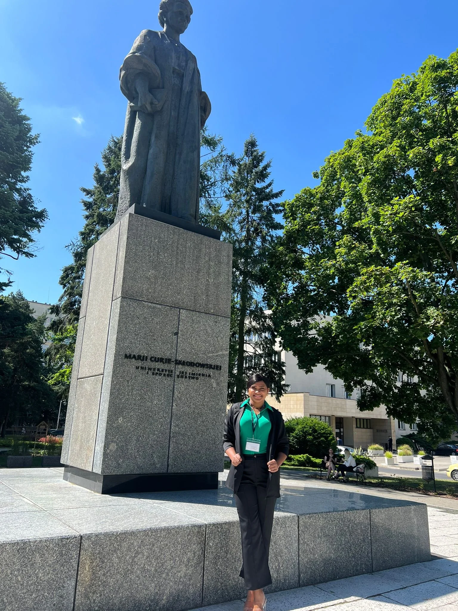 A woman in a teal blouse and black skirt standing next to a tall statue on a large stone pedestal, in an outdoor park with trees and benches, under a clear blue sky.