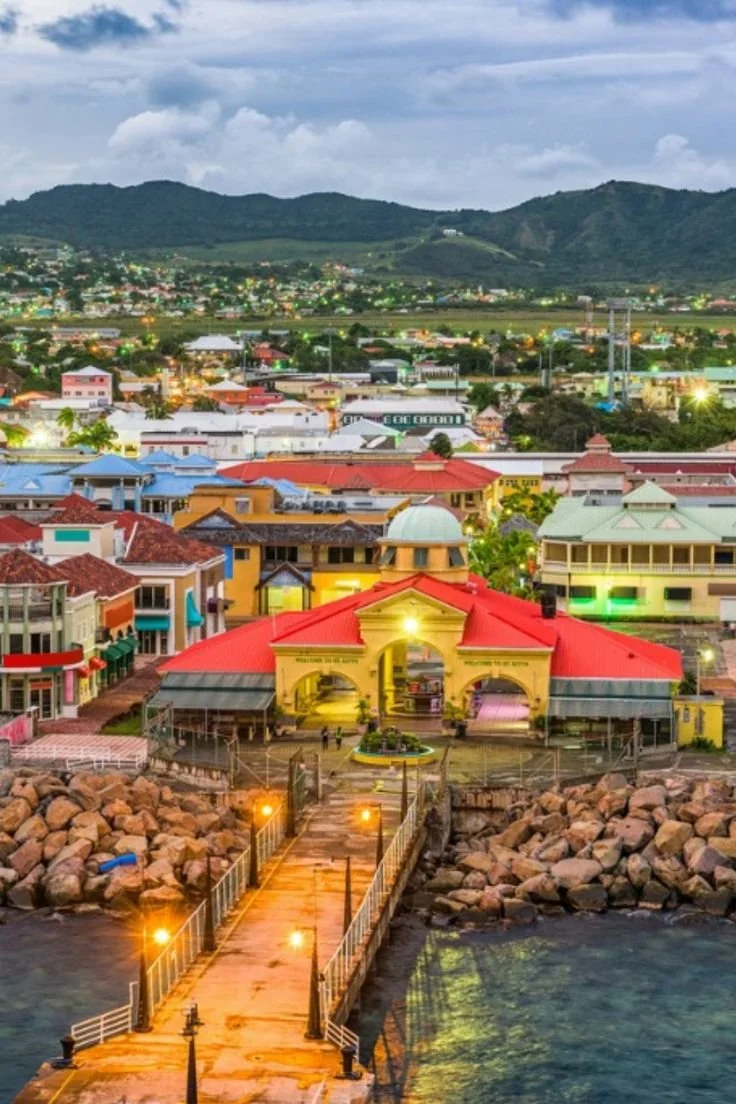 Colorful coastal town with a pier leading to a building with a red roof, surrounded by rocks and water, with hills and a cloudy sky in the background.