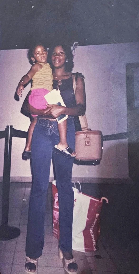 A woman holding a young girl in her arms, standing indoors against a wall. The woman is smiling, wearing a black top and high-waisted jeans, with a beige handbag on her shoulder. The girl is wearing a yellow top and pink shorts, looking away from the camera.