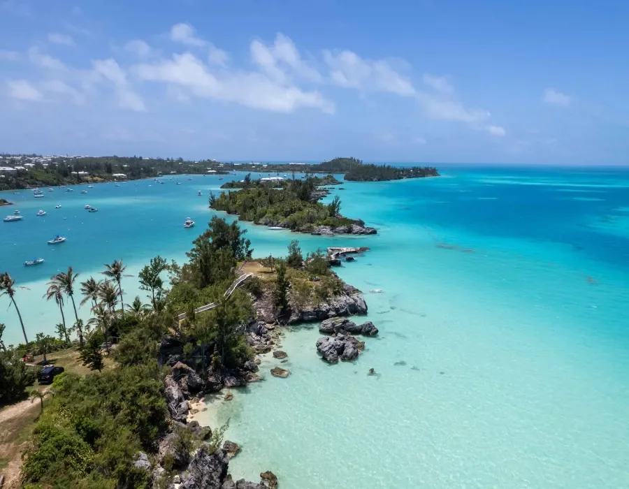 A tropical coastal landscape with turquoise waters, small boats, lush green vegetation, and a mix of sandy and rocky shoreline under a partly cloudy sky.