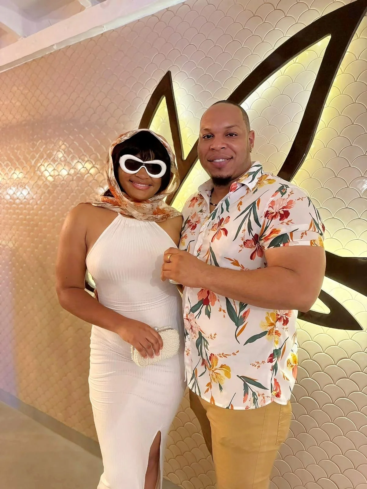 A man and woman posing together indoors. The woman is wearing a white dress, sunglasses, and a headscarf. The man is wearing a floral shirt and beige pants. The background features a decorative wall with a pattern and lighting.