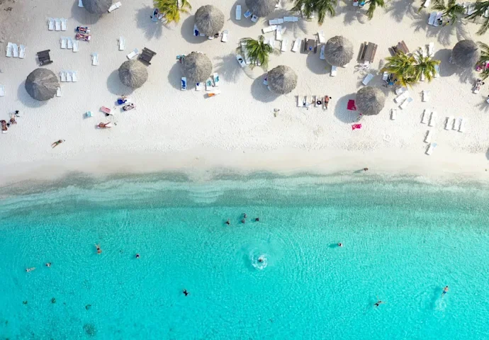Aerial view of a beach with white sand, turquoise water, and people swimming. Beach umbrellas and lounge chairs are set up along the shore, with some palm trees providing shade.